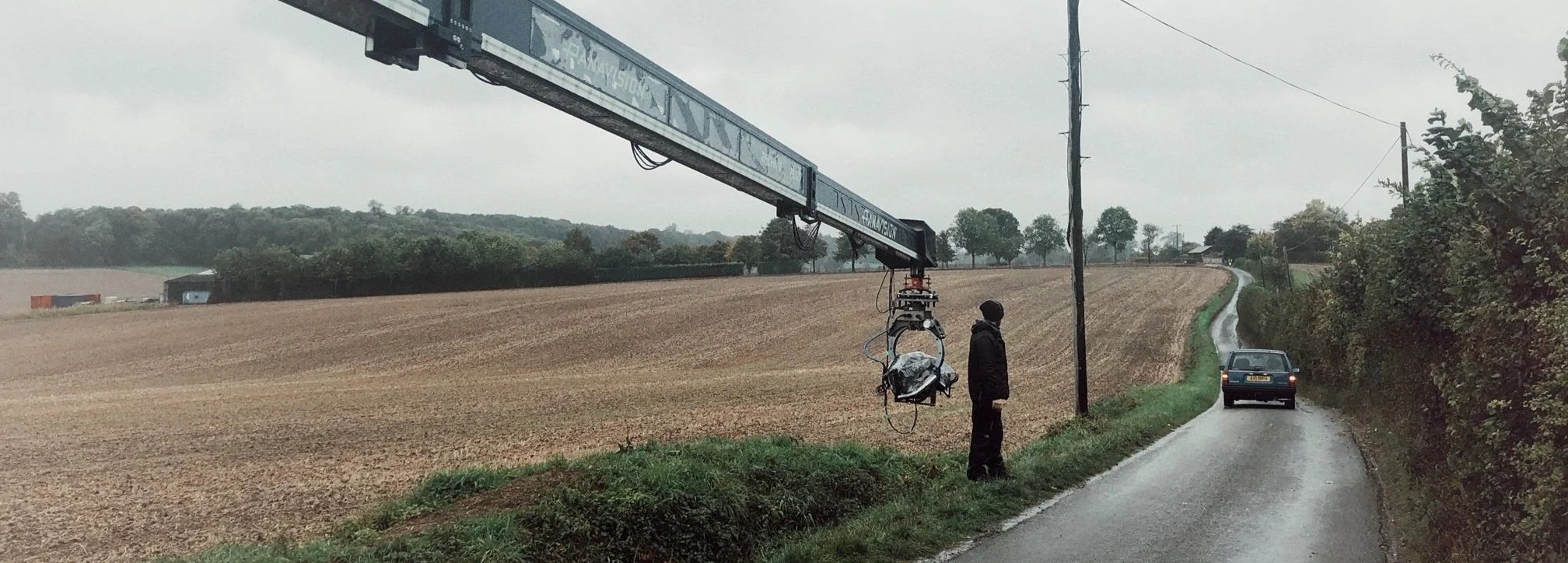 A film crew is filming a scene with a camera mounted on a crane next to a rural road. A man stands near the crane, which is positioned next to a telephone pole. The scene is set in a countryside with fields, trees, and a cloudy sky.