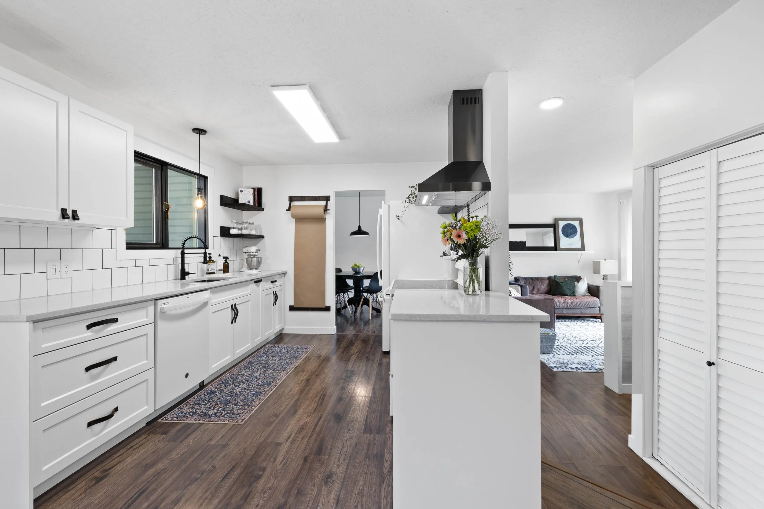 A modern, bright kitchen with white cabinets, black hardware, white subway tile backsplash, dark wood flooring, and a small window above the sink. There is a vase of colorful flowers on the white countertop. In the background, a dining area with black chairs and a round table, a white refrigerator, and a glimpse of the living room with a sofa and framed pictures on the wall.