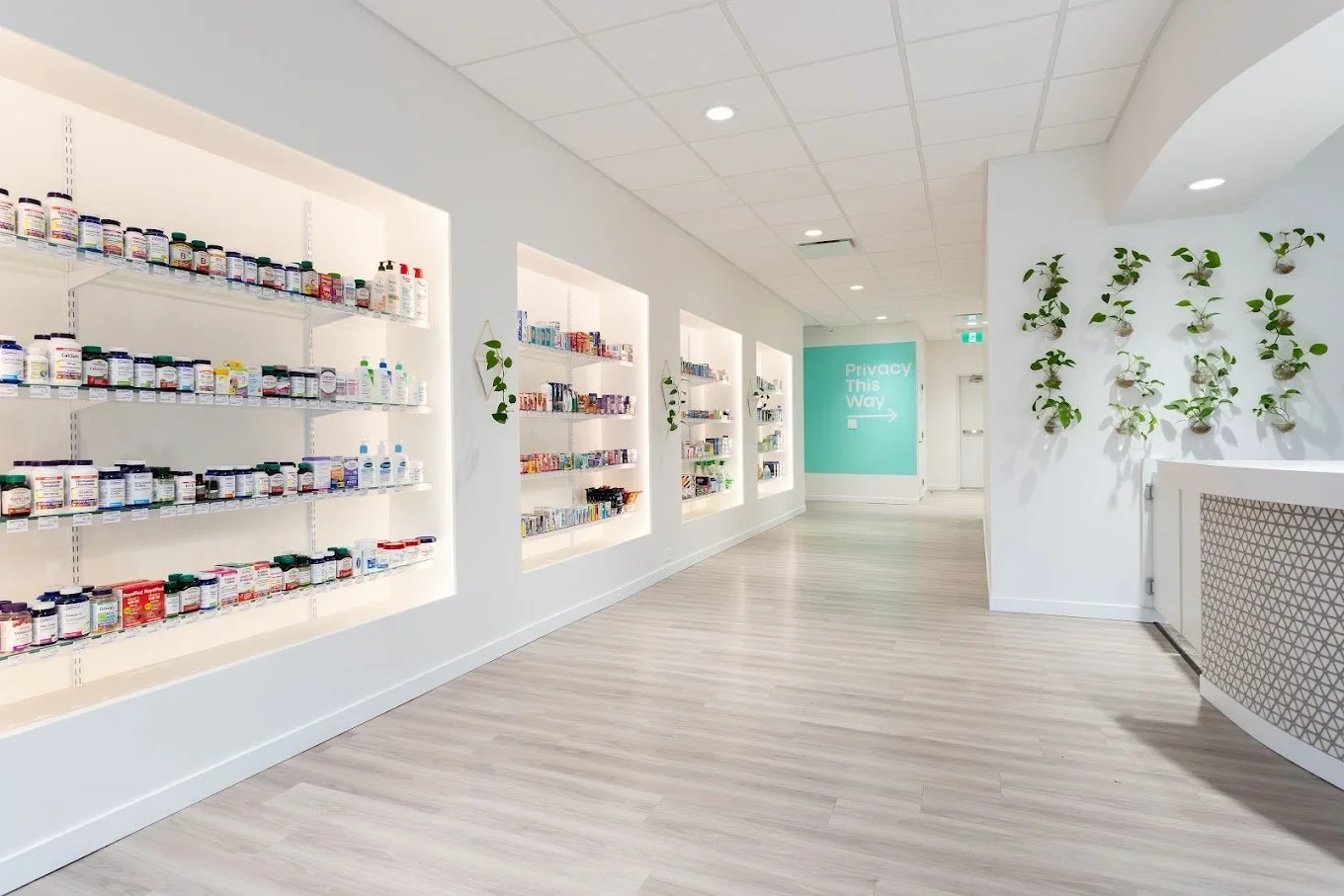 Interior of a bright and modern pharmacy with white shelves filled with medication bottles and boxes, light wood flooring, and green plants on the walls. Commercial tenant improvement project.