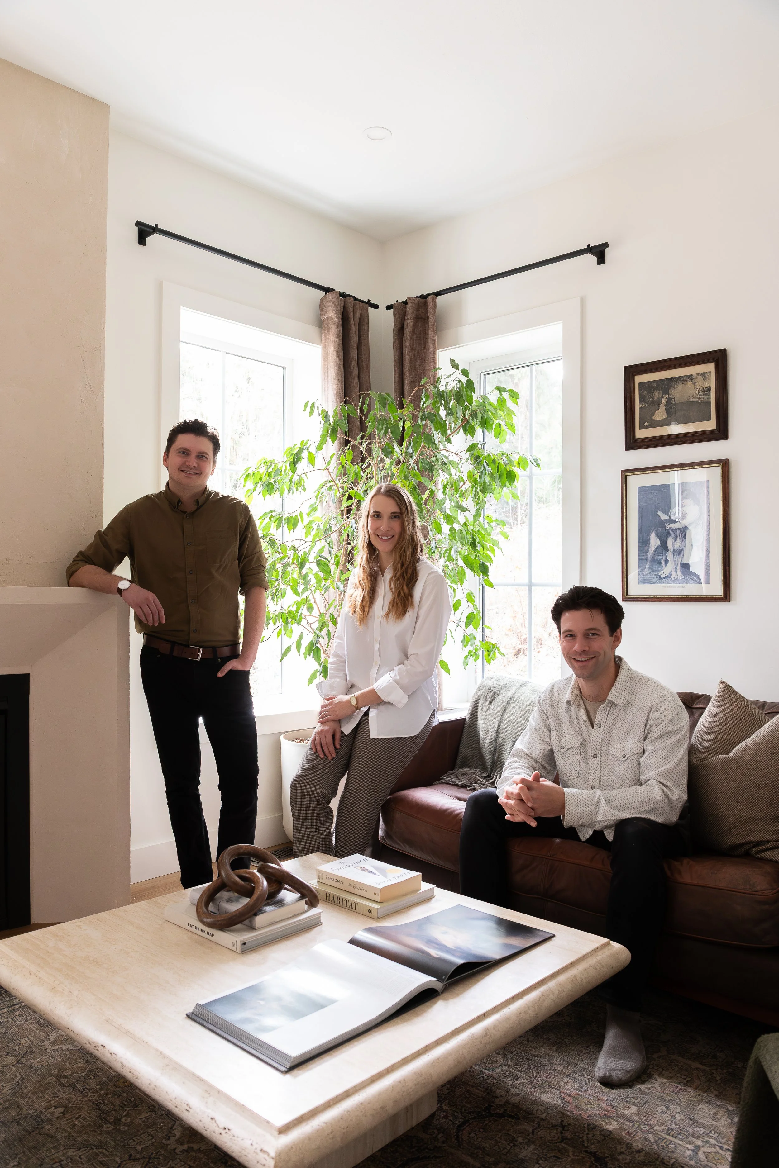 Three people in a living room with a weeping fig, vintage pictures, and books on a travertine coffee table, smiling at the camera.