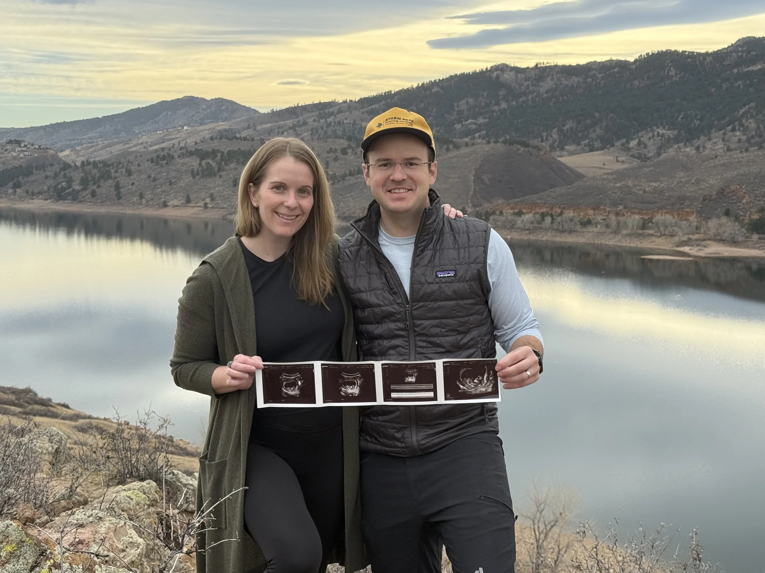 A smiling woman and man holding ultrasound pictures in front of a lake with mountains in the background at sunset.