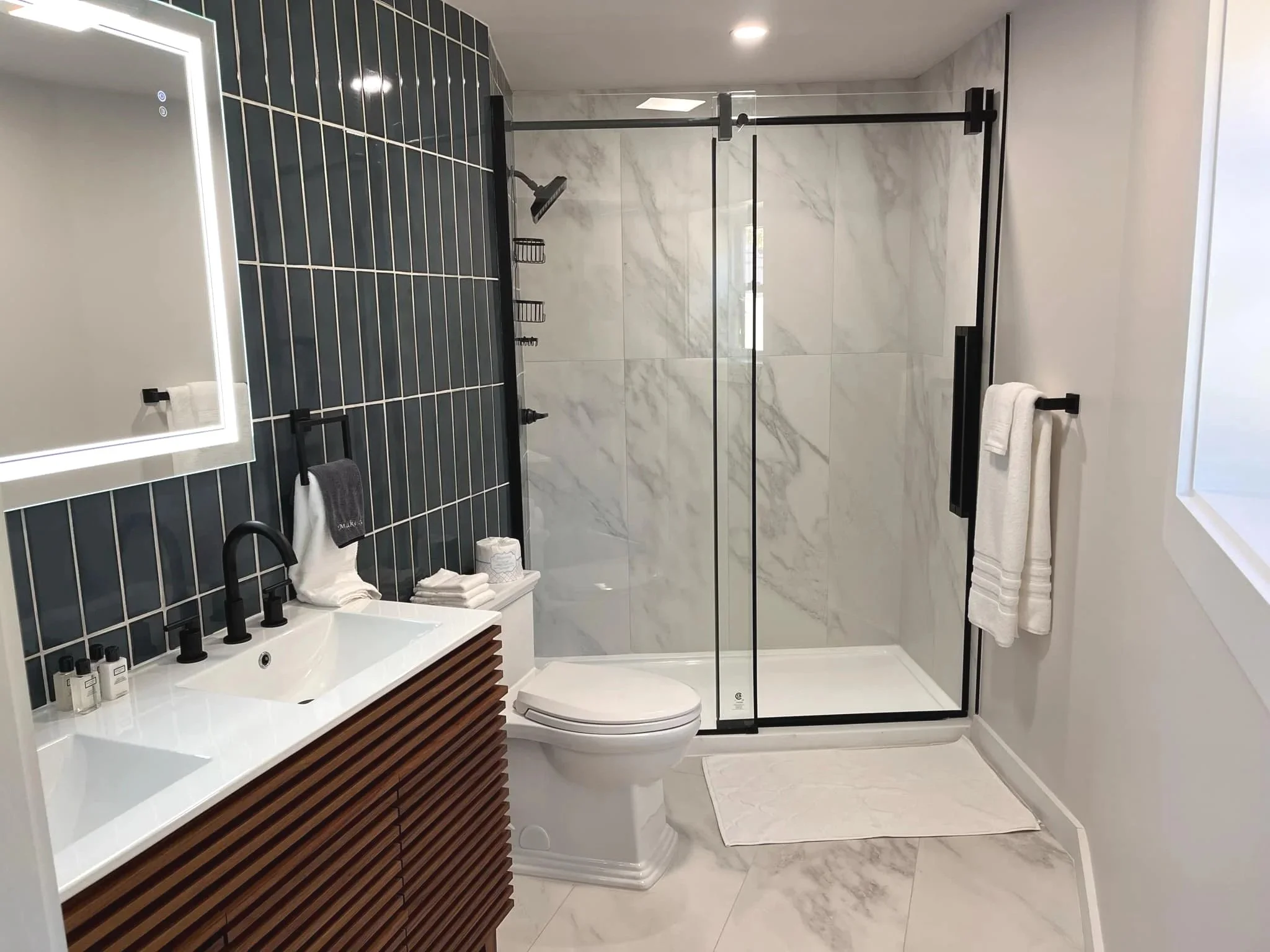 Modern bathroom with black-tiled wall, white marble shower, and wooden vanity with black fixtures, white towels, and a mirror with LED lighting.