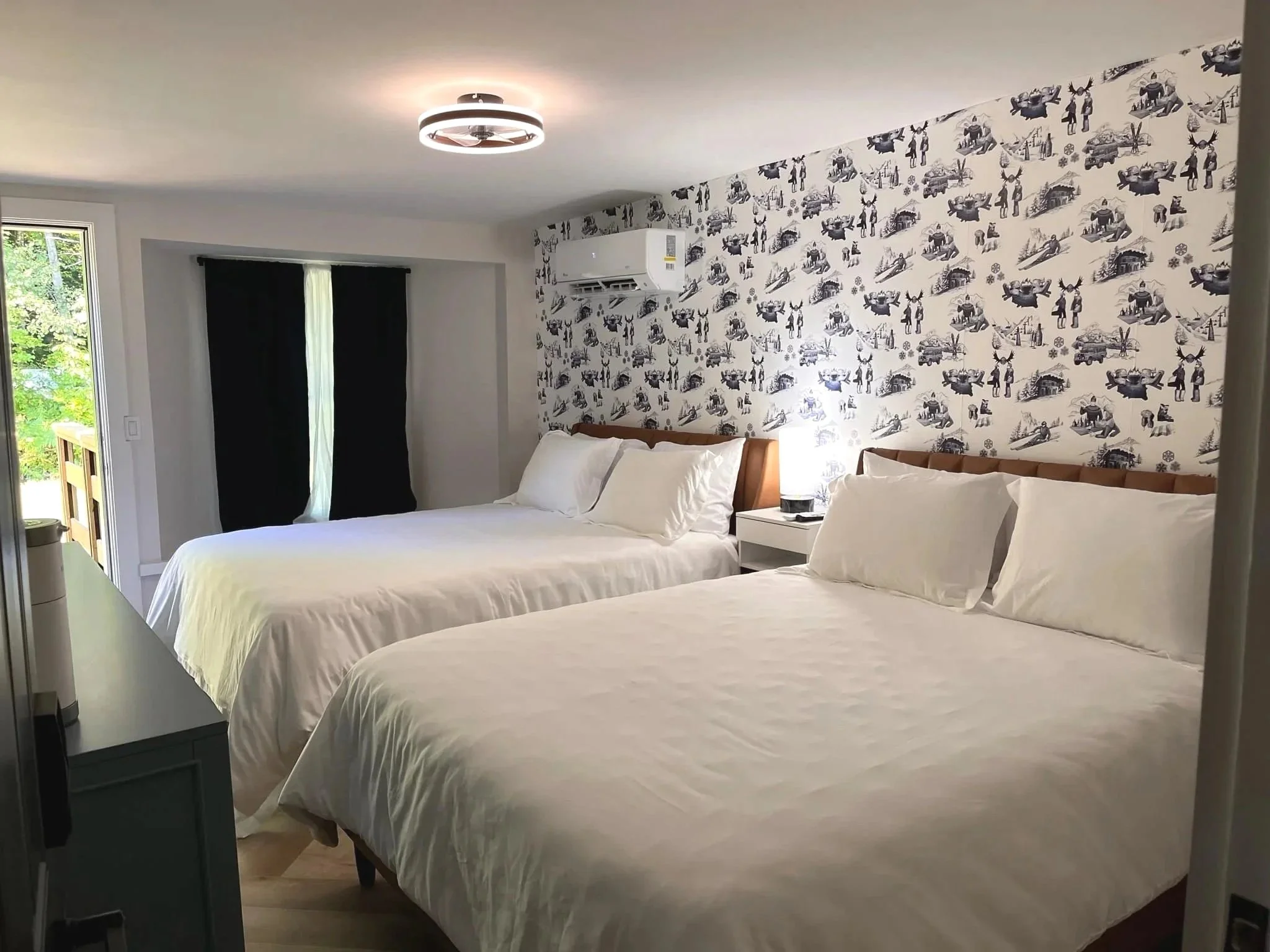 Two beds with white linens and pillows in a bedroom with a patterned wallpaper featuring black silhouettes of animals, trees, and people, with a window and a sliding door with black curtains on the left side.