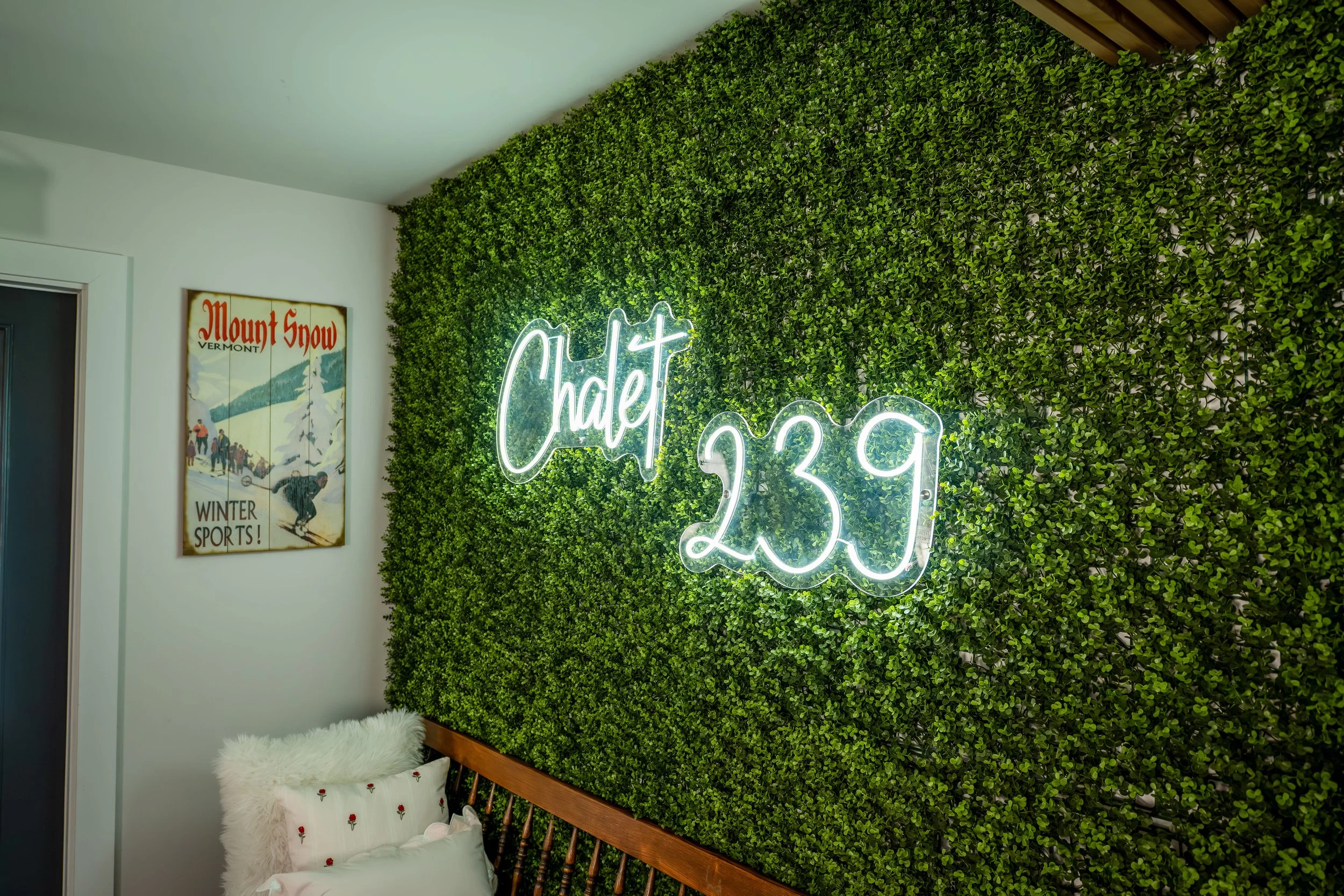 Interior of a room featuring a green leafy wall with a neon sign reading 'Chalet 239'. To the left, there's a framed vintage Mount Snow Vermont Winter Sports poster, and a bed with white furry pillow and decorative pillows.