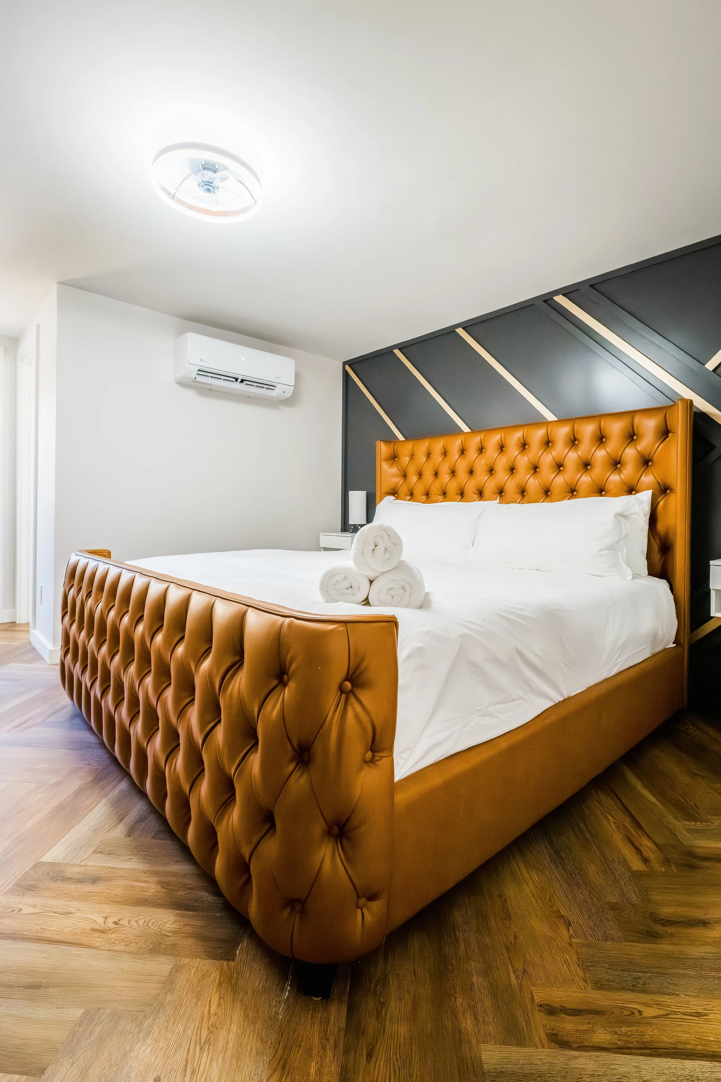 A bedroom with a leather tufted bed, white bedding, and three rolled towels on top. The headboard is orange-brown leather, and there are white pillows behind the towels. There is a wall with a decorative black and gray pattern behind the bed, a night