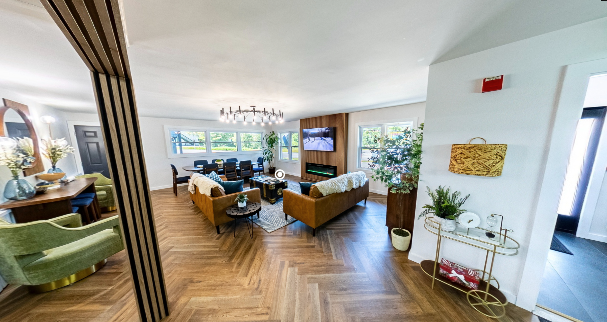 A modern, bright living room with a wooden floor, a comfortable seating area with two brown sofas, a wall-mounted TV, and large windows letting in natural light.