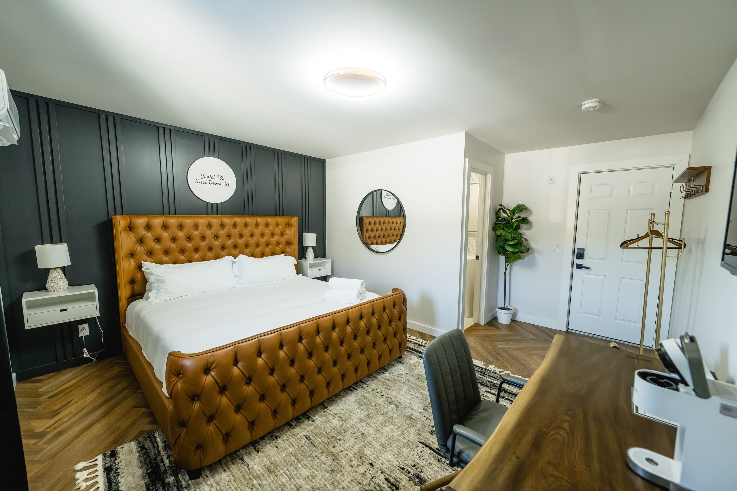 Hotel room with a large bed featuring a tan tufted headboard, white bedding, two nightstands with lamps, a black accent wall behind the bed, a round mirror on the white wall, a potted plant near the door, a wooden desk with a black chair, a coat rack