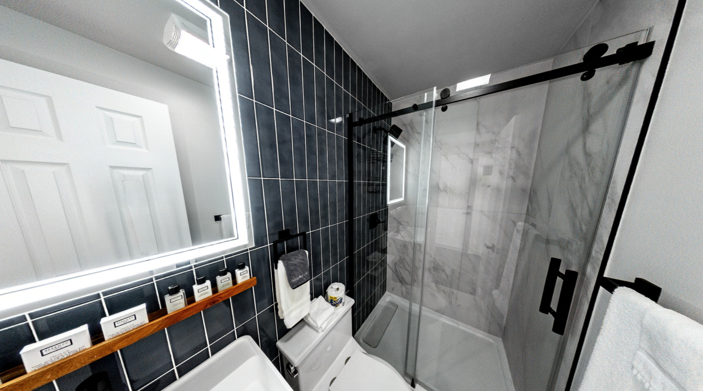 A modern bathroom with black tiled walls, a glass shower enclosure with marble interior, white fixtures, a rectangular mirror with a backlit frame, and a wooden shelf holding toiletries. There are towels hanging on a black towel holder and a window i