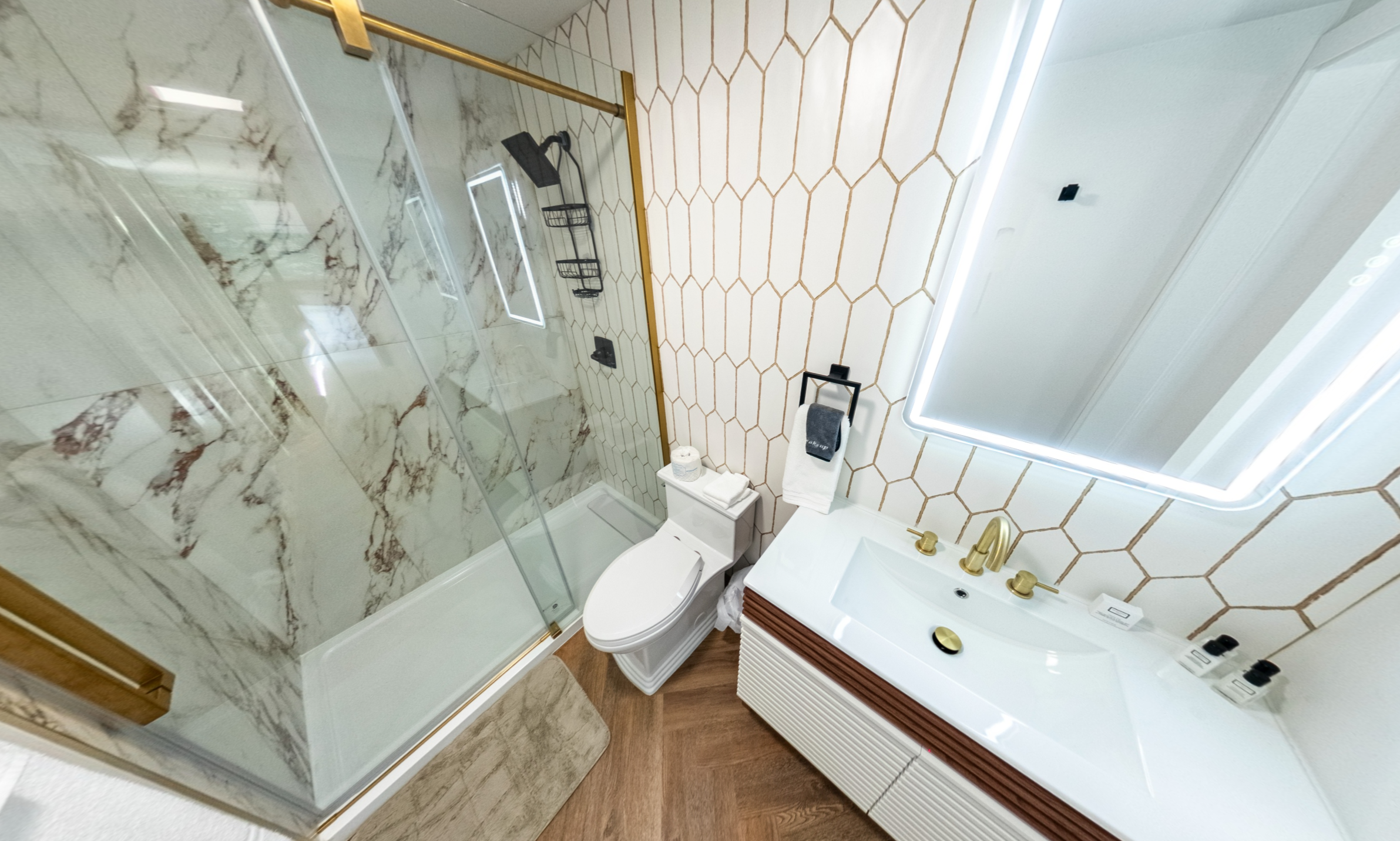 Bathroom with a walk-in shower with marble walls and a glass door, a white toilet, a vanity with a white sink and gold fixtures, a large mirror with LED lighting, black towel and shelf accessories, and hexagon patterned wall tiles.