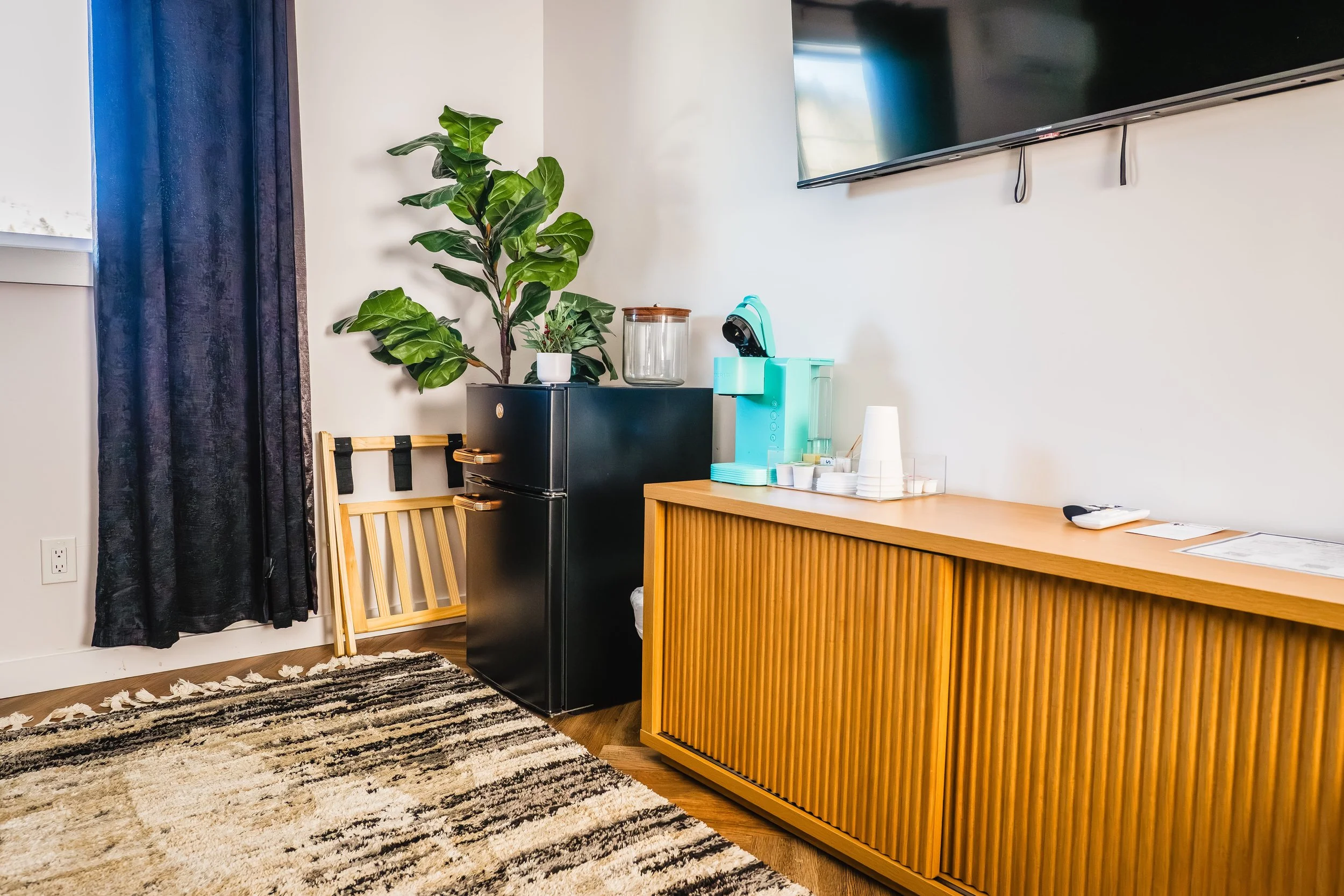A corner of a room featuring a large flat-screen TV mounted on the wall, a black mini fridge with a plant on top, a wooden sideboard with a teal coffee machine, disposable cups, and a remote control, a black and white patterned rug, and dark curtains