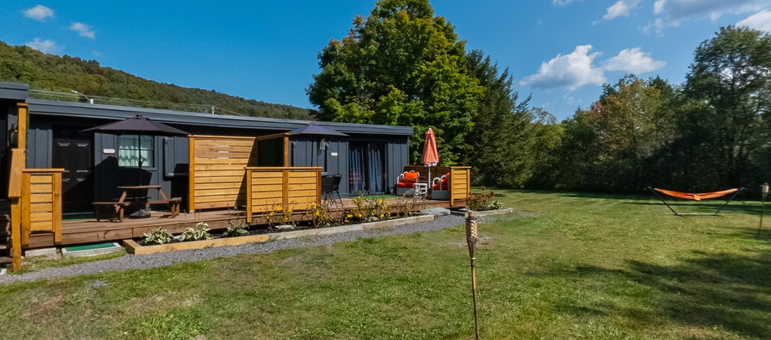 A backyard area with a wooden deck, outdoor furniture, and a large grassy lawn. The deck has patio cushions, an umbrella, and garden beds along its edge. There is a hammock on the lawn, surrounded by trees and a hill in the background under a partly 