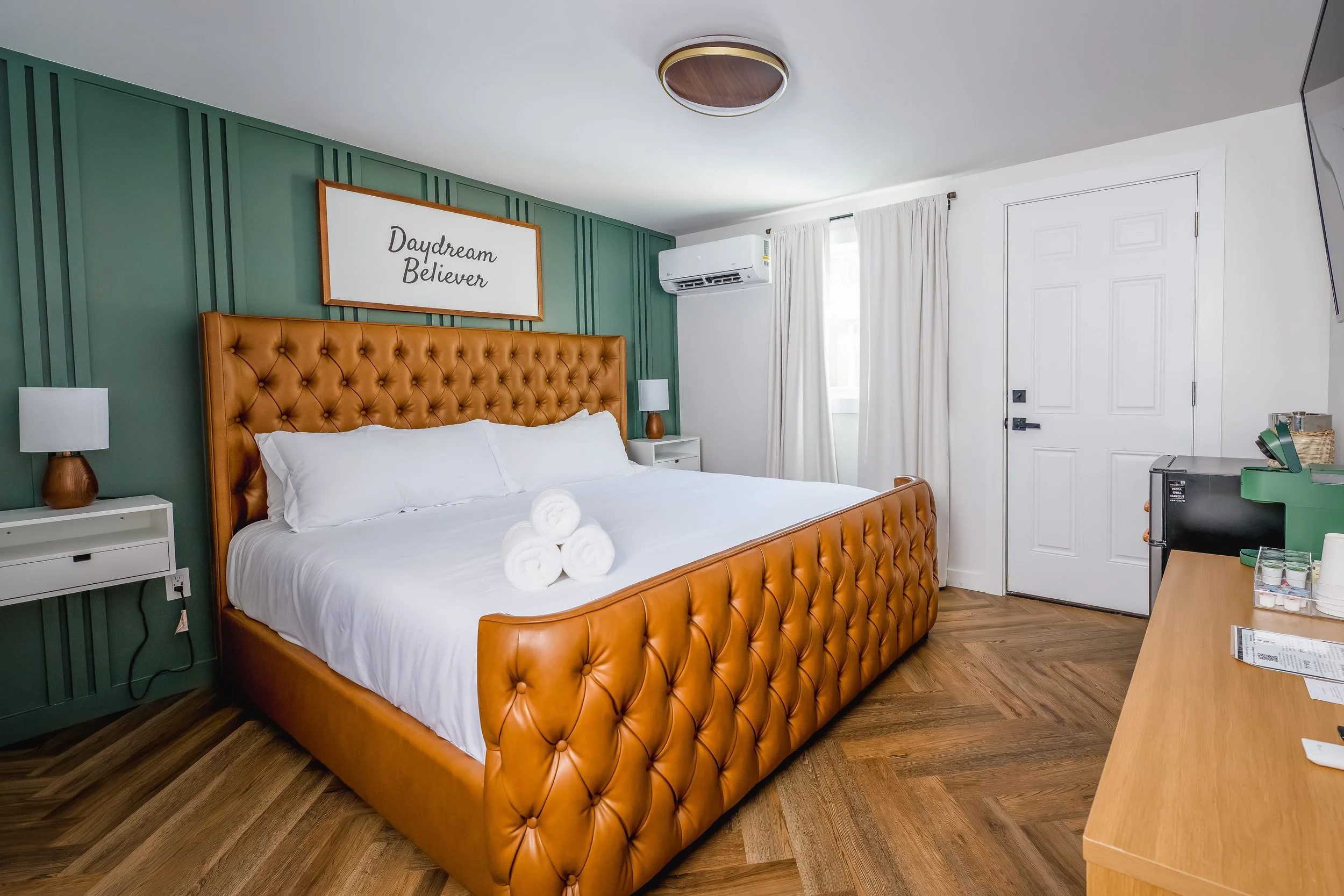 A hotel room featuring a large bed with a tufted leather headboard, white bedding, and rolled white towels. There are two small white nightstands with lamps, green accent wall behind the bed with a framed sign that says "Daydream Believer," a window 