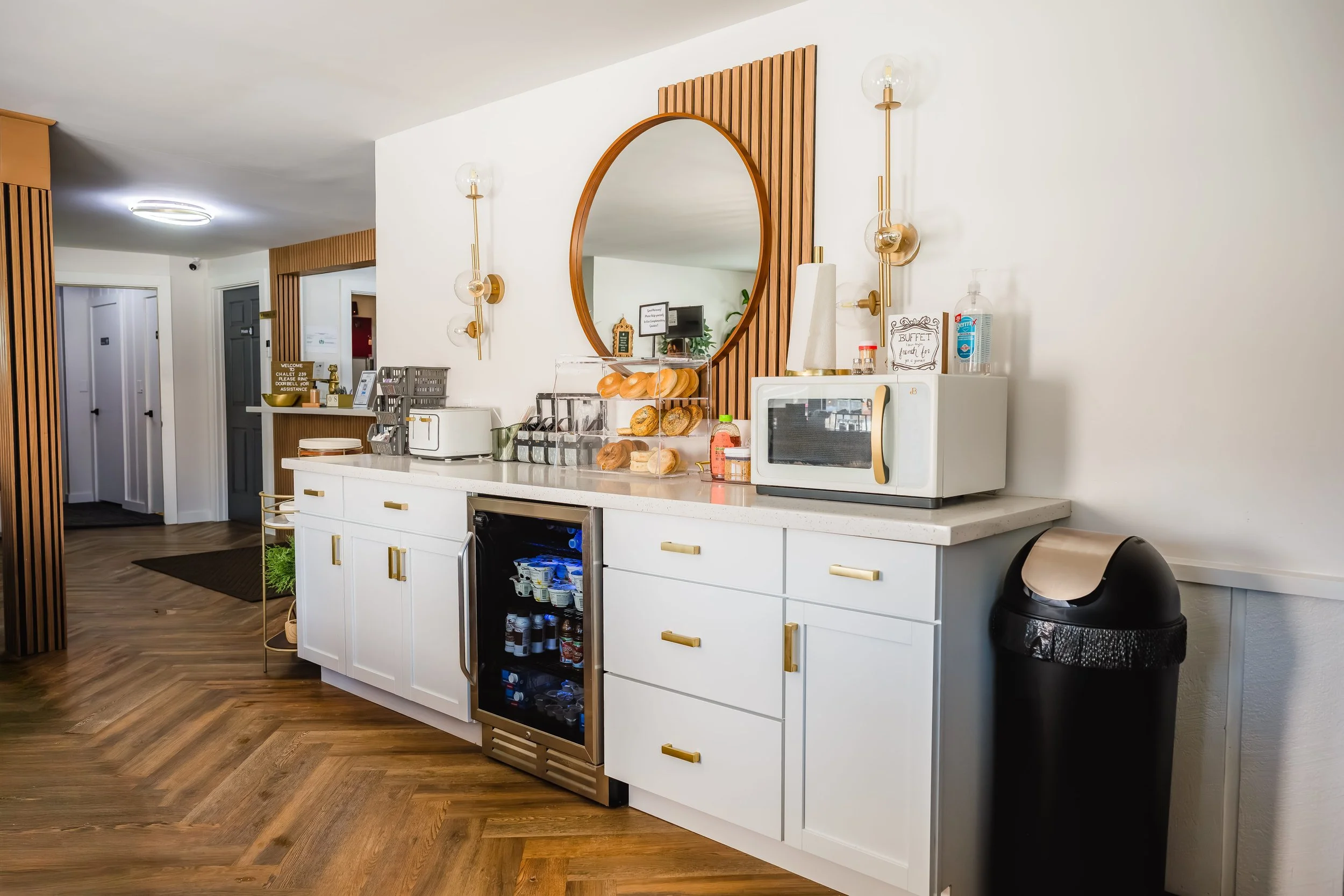 A kitchenette area with a white counter, a small wine fridge, a microwave, and a black trash bin. There is a round mirror on the wall, and various items including bread, jams, and a hand sanitizer on the counter. The floor is wood-patterned.