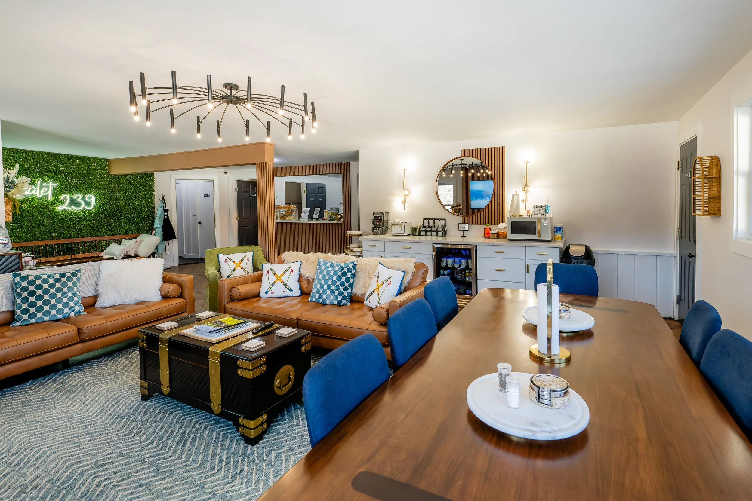 Stylish hotel lounge with leather sofas, colorful pillows, a wooden coffee table, a dining table with blue chairs, modern lighting, a green photo wall, and a kitchenette with a mini-fridge and microwave.