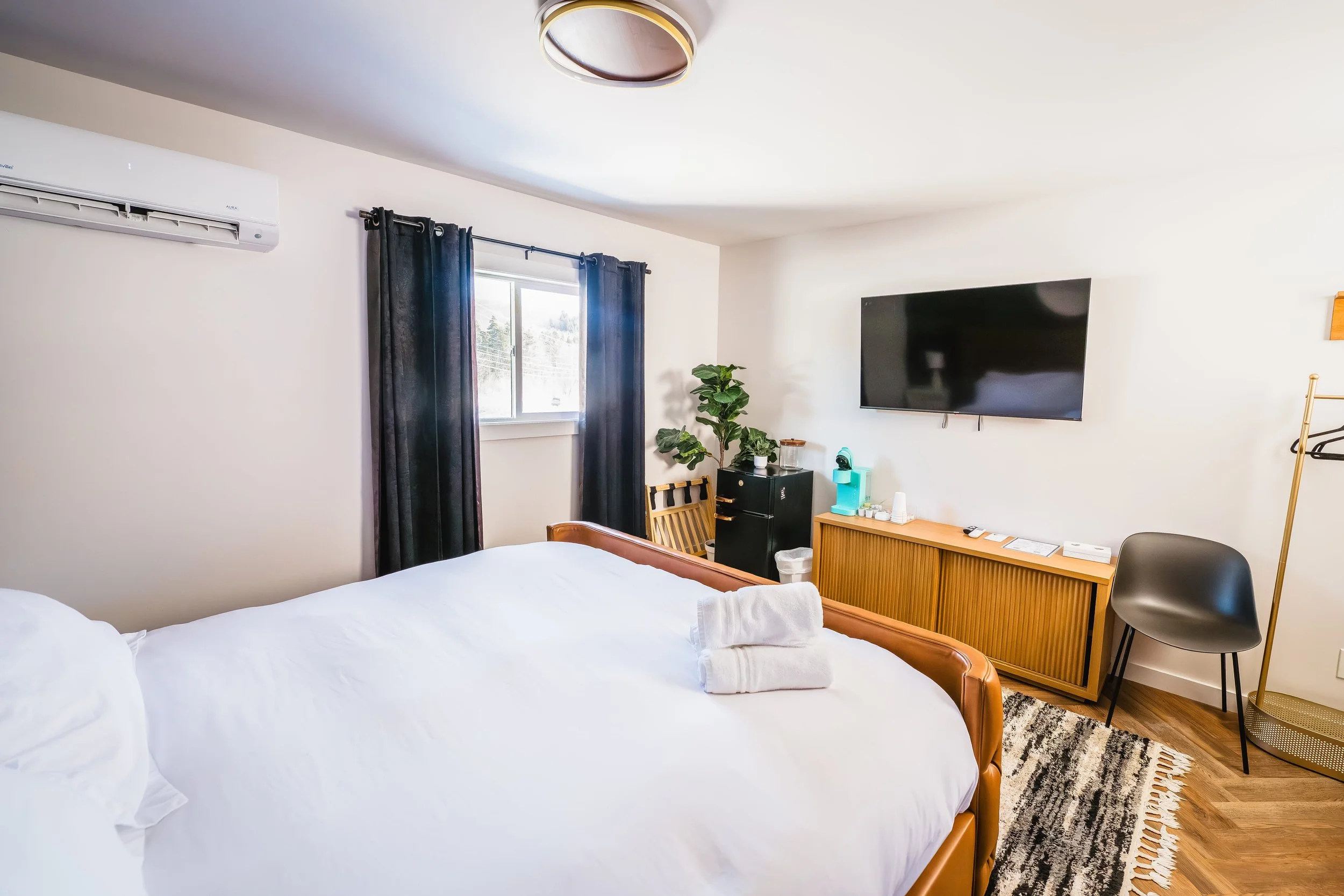 A hotel room with a large bed, black curtains, a window, a TV mounted on the wall, a wooden dresser, a black chair, a potted plant, and various amenities on the dresser.