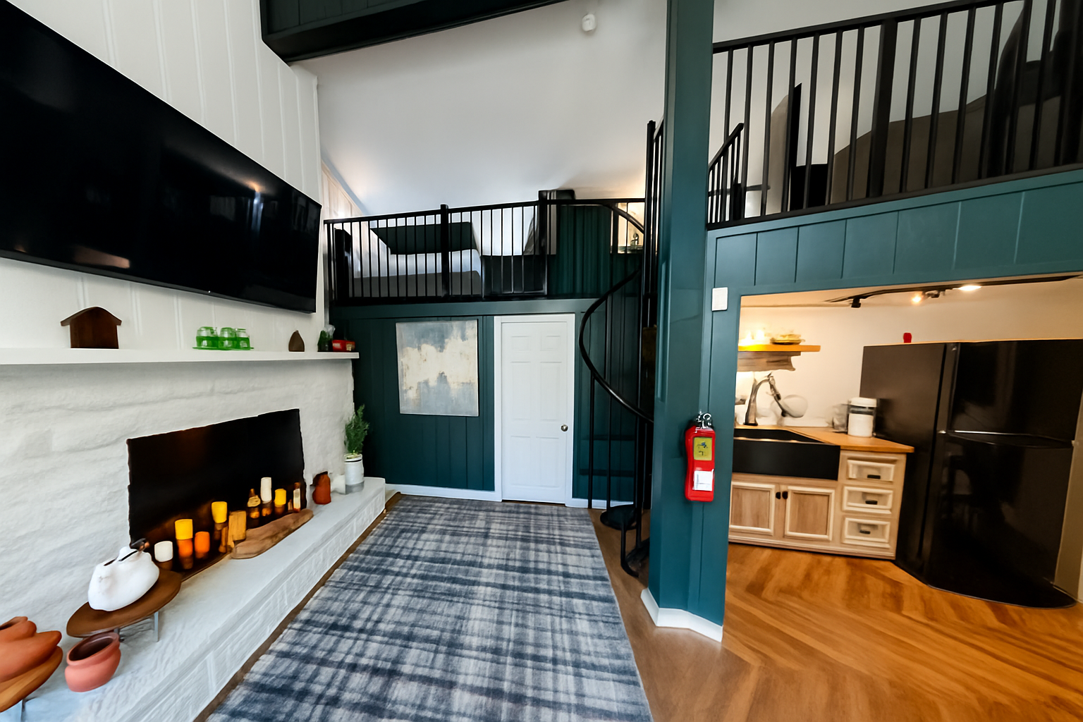 Living room with white fireplace, television, and decorative candles, open kitchen with black refrigerator and wooden cabinets, staircase leading to loft area, green walls, hardwood flooring, and various decorative items.