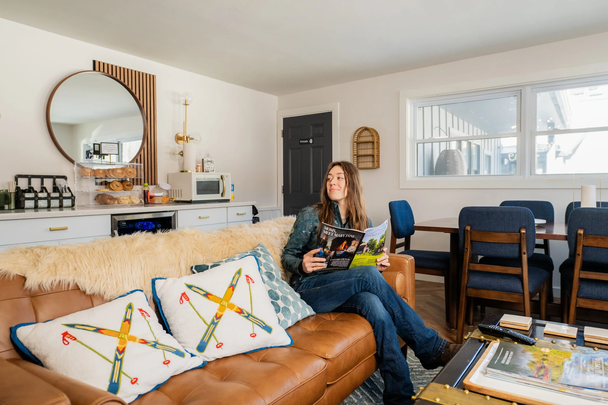 Woman sitting on a tan leather sofa with decorative pillows, reading a magazine in a modern living room with a dining area and decorated with a large round mirror and window.