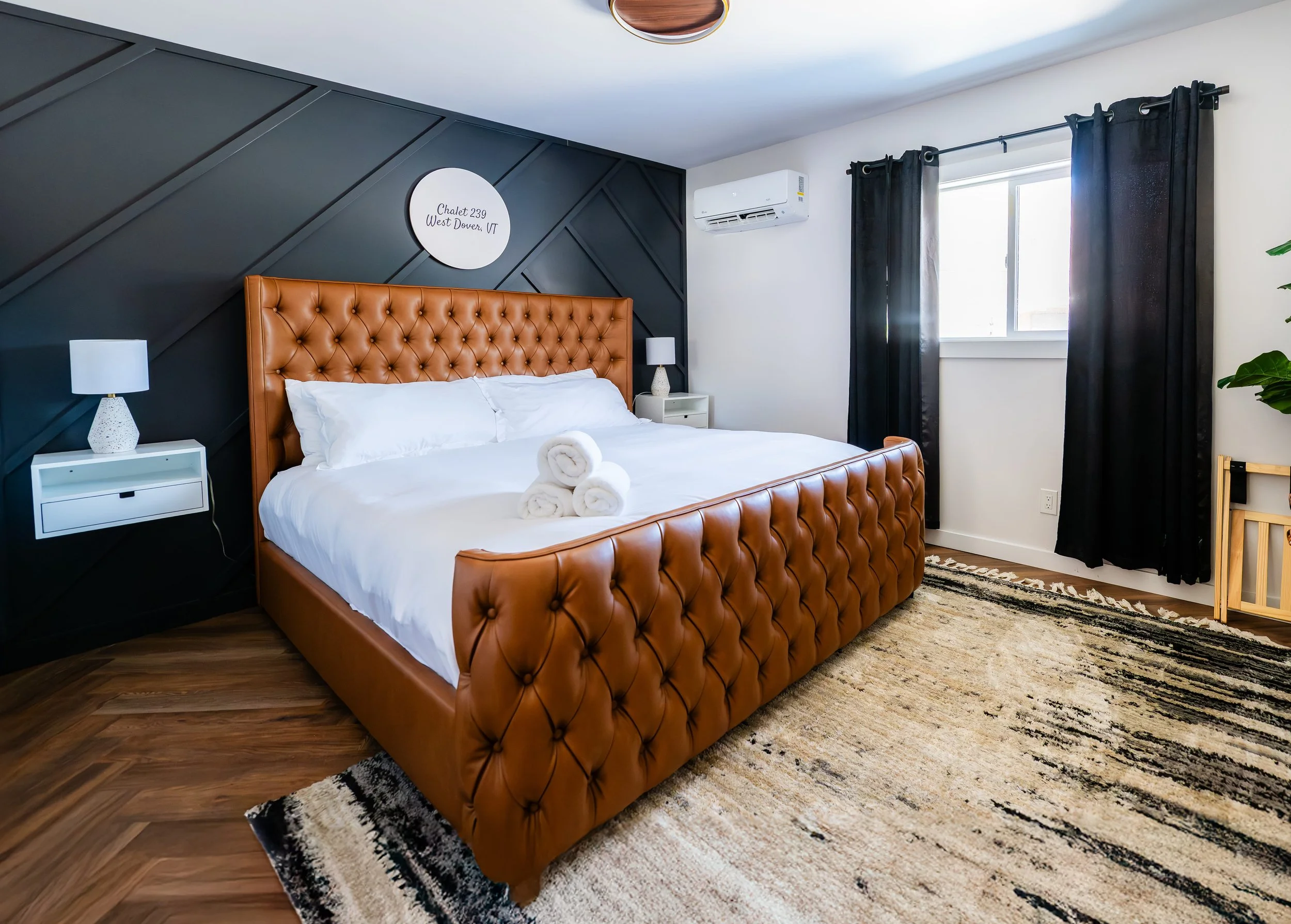 A bedroom with a large leather-upholstered bed, white bedding, and rolled towels on top. Dark accent wall behind the bed with a white circular sign reading "Chalet 239 West Dover VT." Black curtains on a window, a white air conditioning unit, and a b