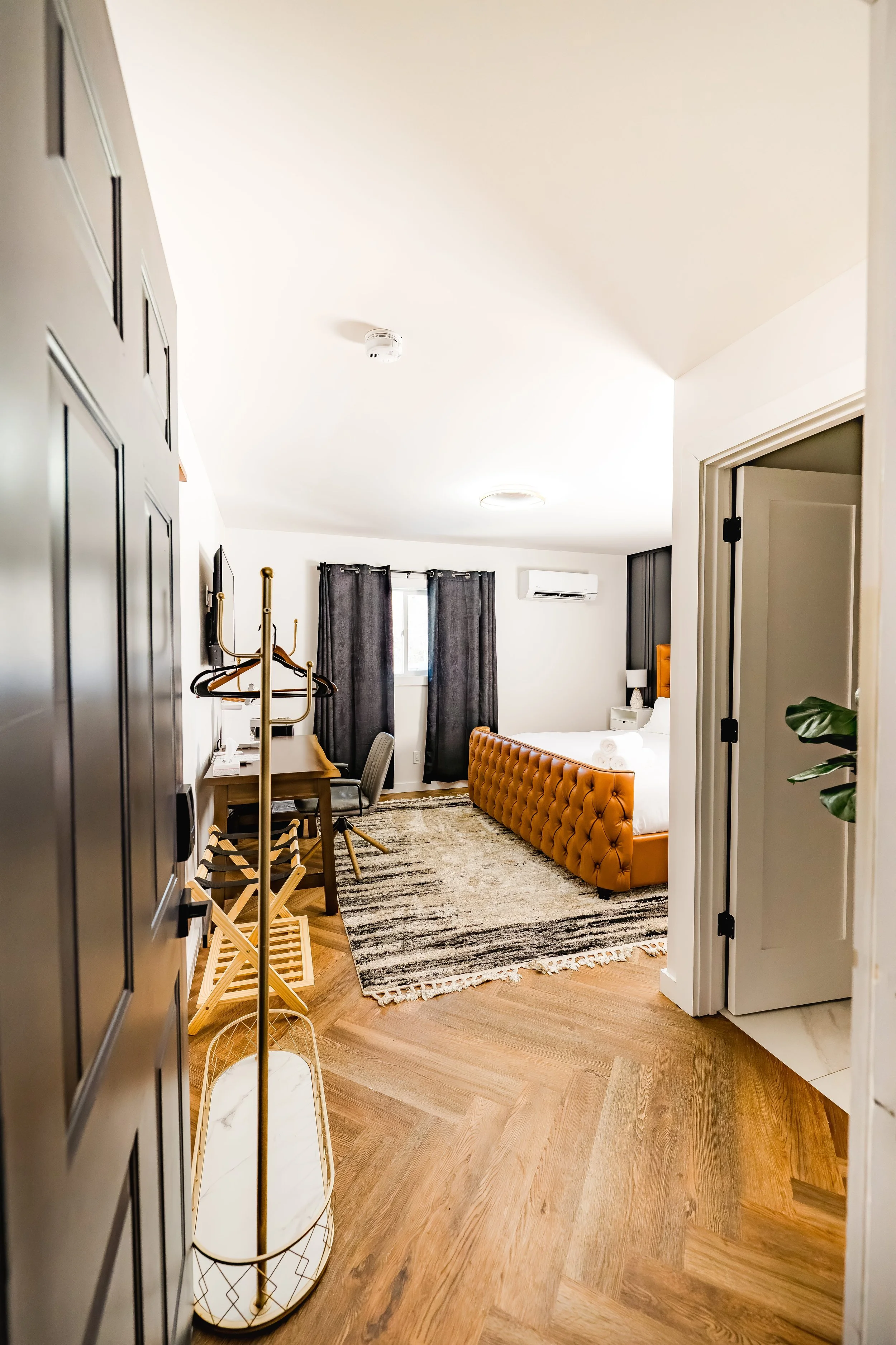 A hotel room featuring a bed with a tufted orange headboard, dark curtains, a desk with a chair, and a closet with a gold clothes rack inside. The room has a wooden floor and white walls.