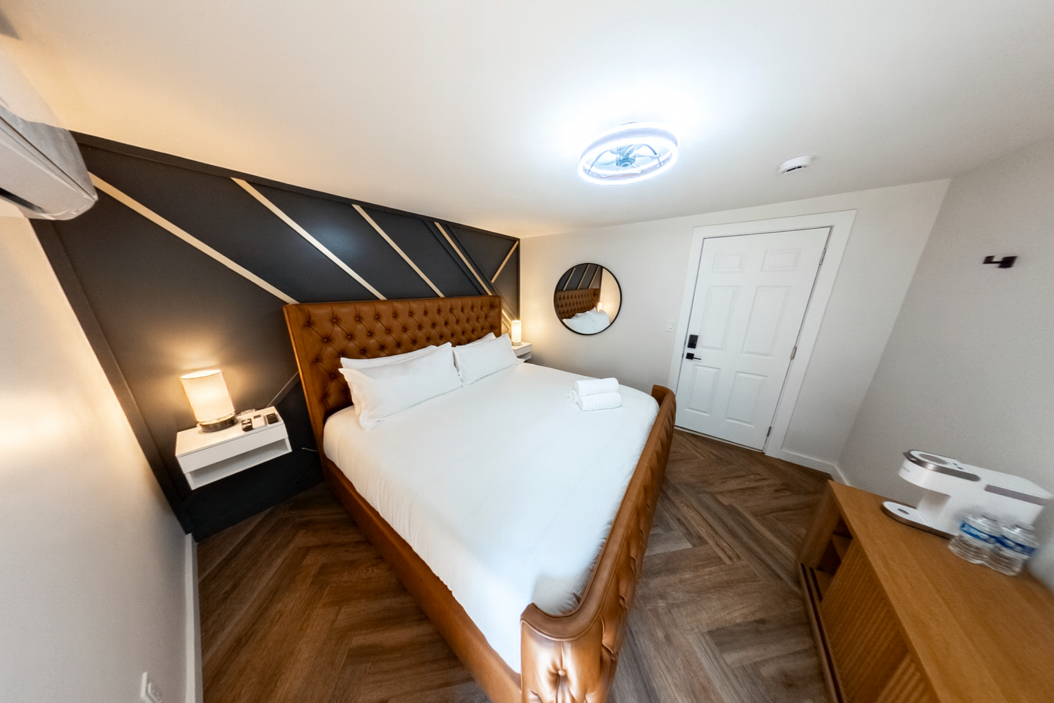 A modern hotel room with a large bed, tufted leather headboard, dark accent wall, and wooden flooring. There are bedside lamps, a mirror, a table with bottled water, and a ceiling light fixture.