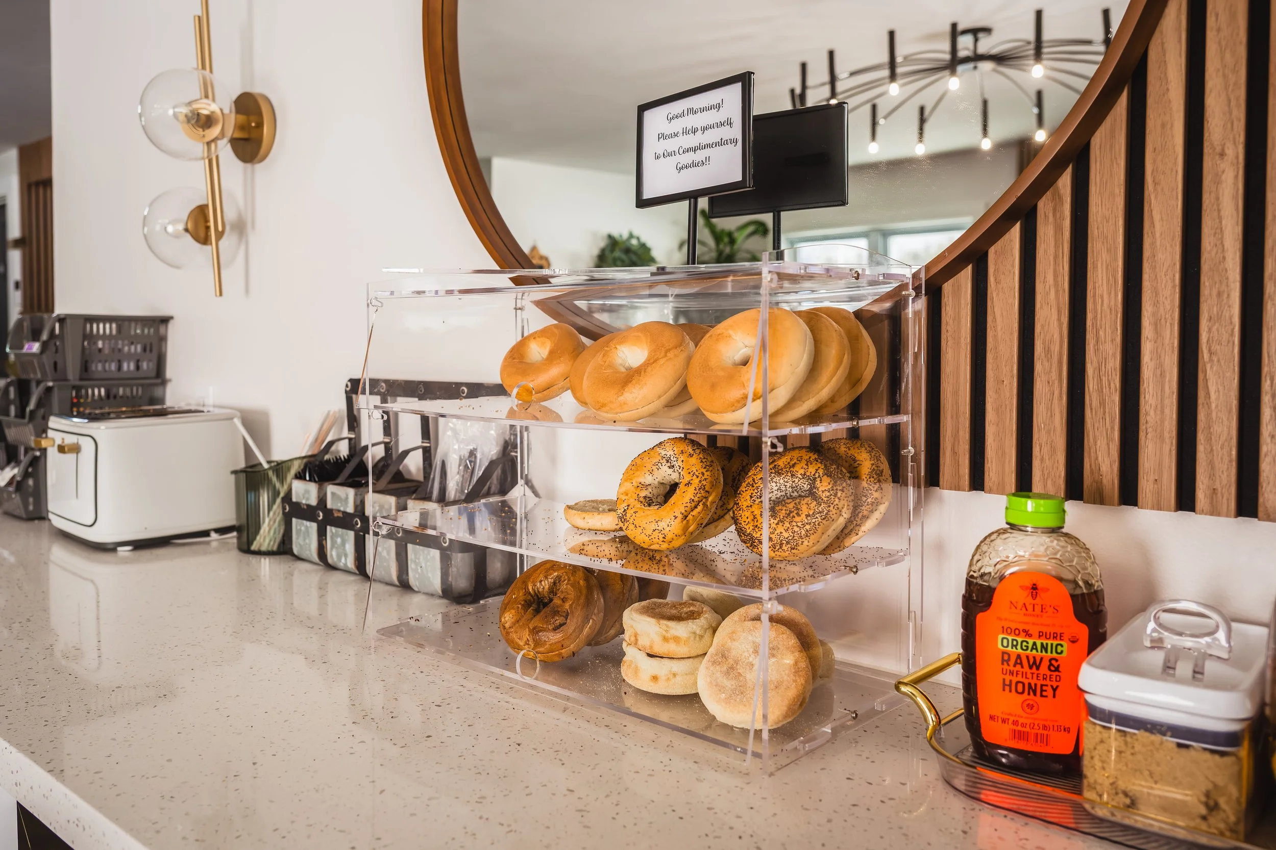 A clear acrylic display case with assorted bagels on a kitchen counter, including plain, poppy seed, and cinnamon raisin varieties. Behind the display, a round mirror hangs on the wall, with a sign above the display offering a morning greeting, and a