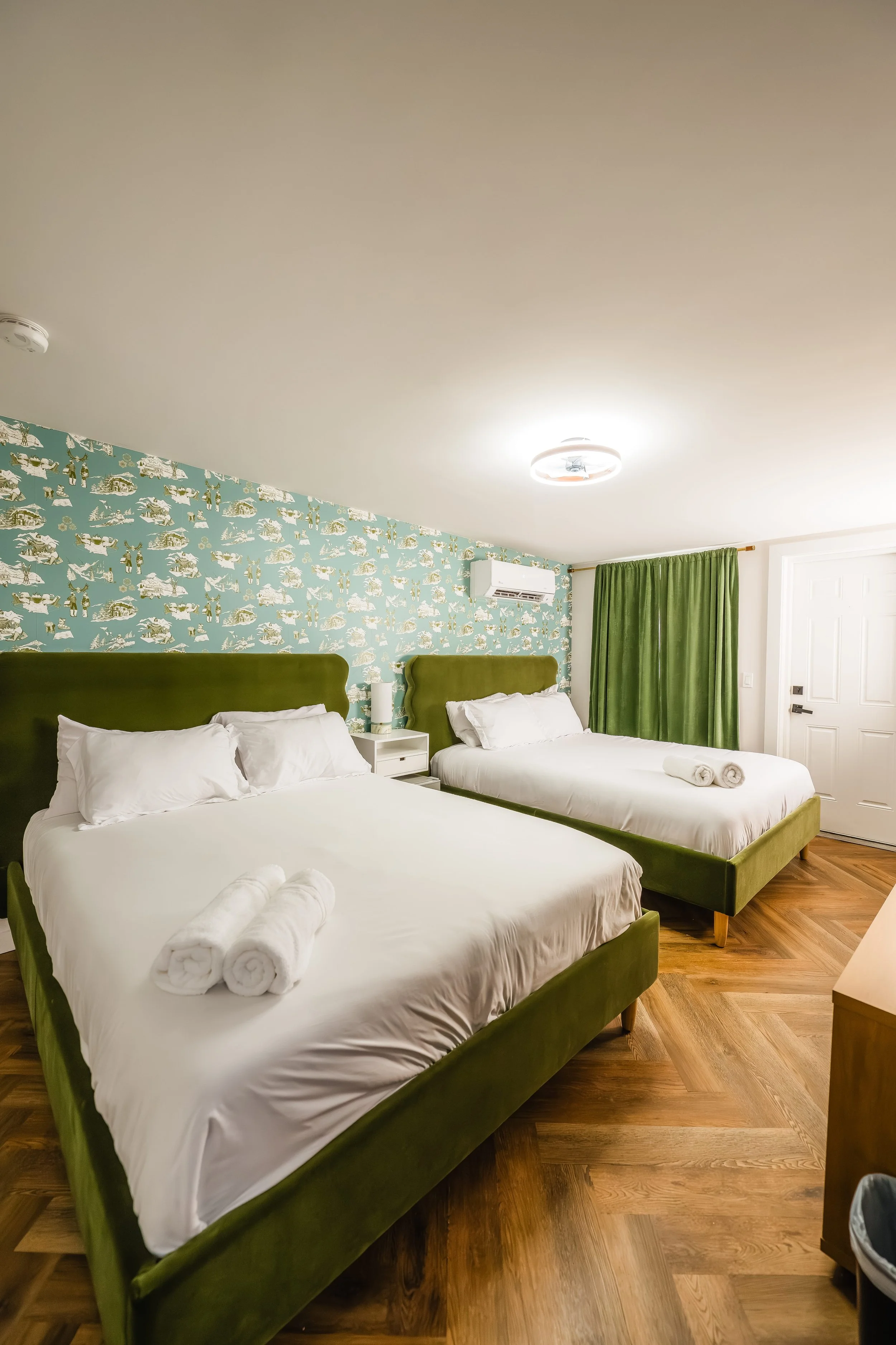 Hotel room with two green beds, white linens, rolled towels, patterned wallpaper, green curtains, and hardwood floors.