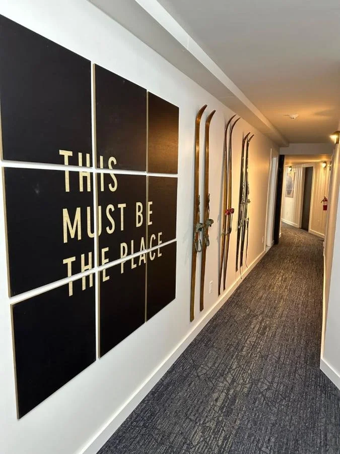 Hallway with wooden skis mounted on the wall and a black sign that says 'This Must Be The Place' in white letters.