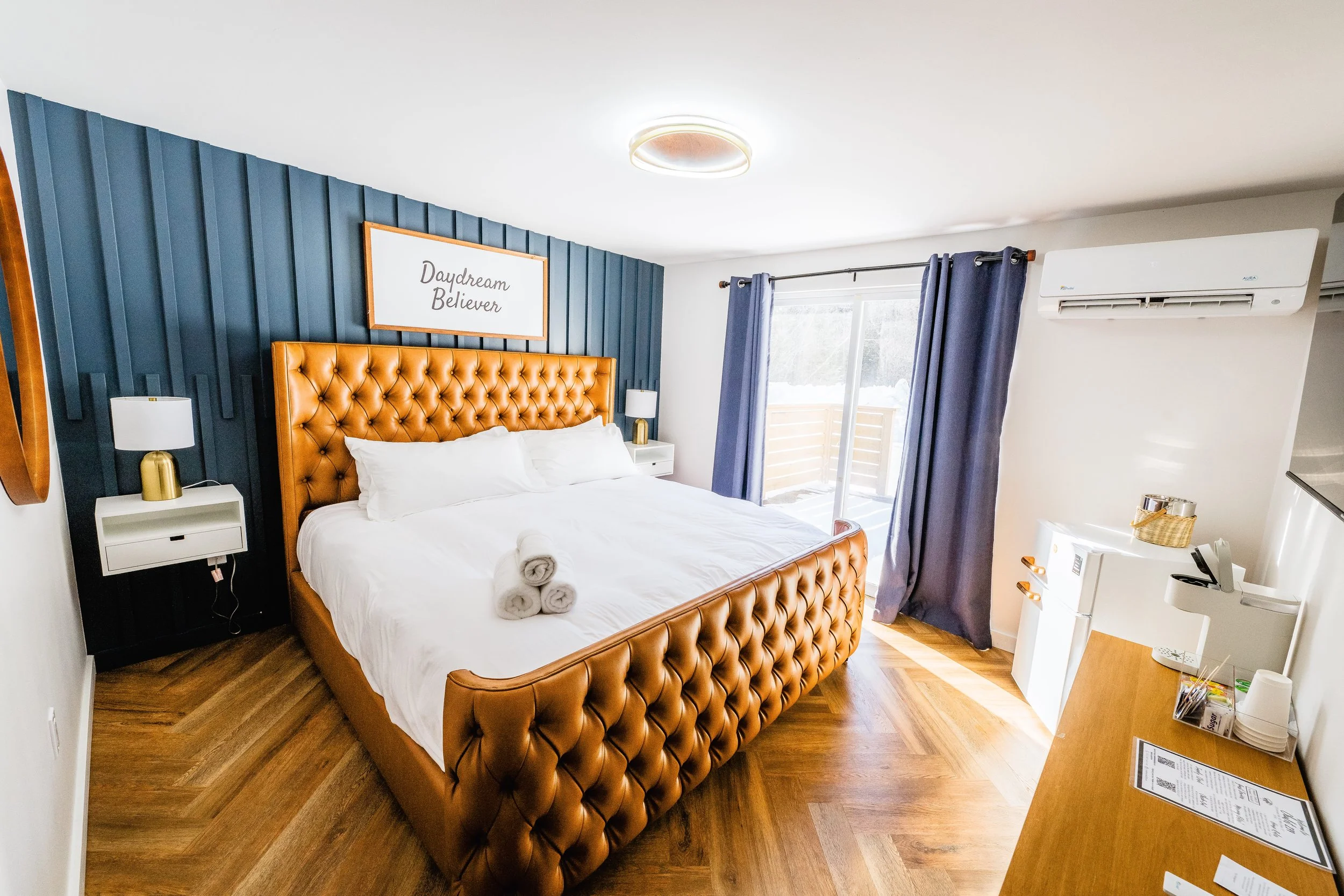 Hotel room with a large bed, tufted leather headboard, blue accent wall, white and gold lamps, towels on the bed, sliding glass door with blue curtains, wooden floor, mini fridge, air conditioning unit, and a wooden desk.