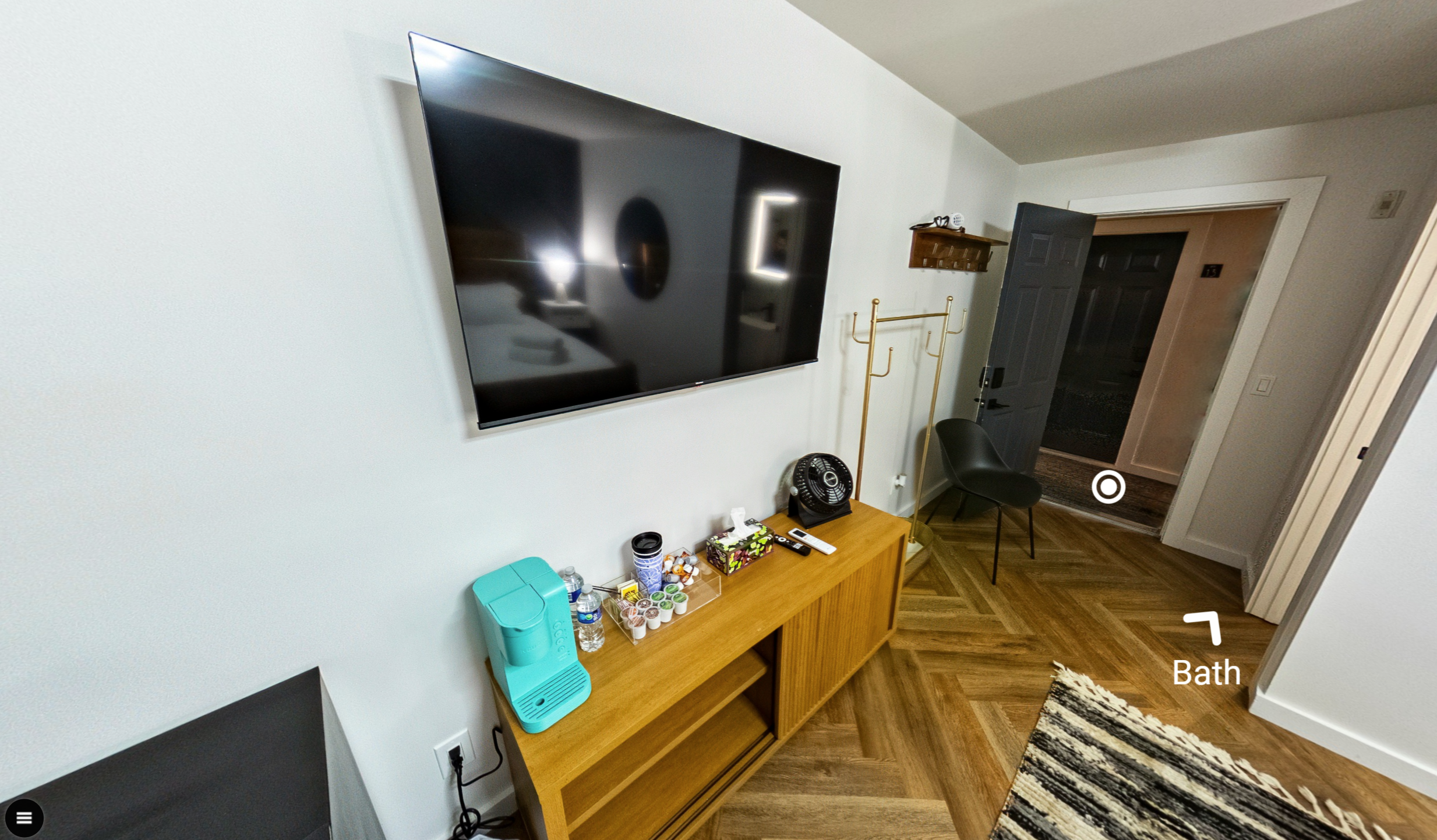 Living room with mounted flat-screen TV, wooden sideboard with coffee pods, water bottles, and a small turquoise water dispenser, black chair, coat rack, and open door leading to an entryway with a rug on wooden floor.