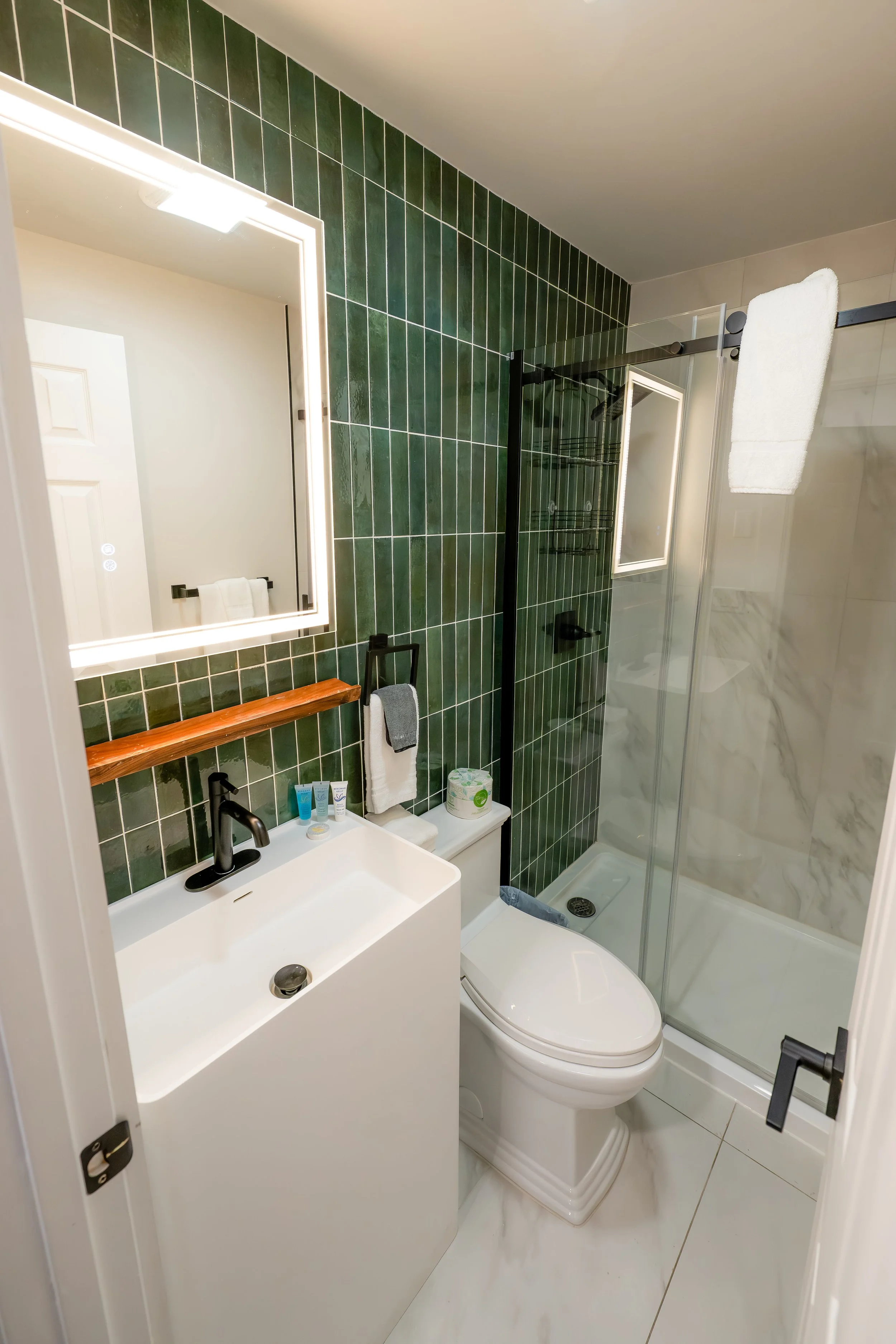 Small modern bathroom with dark green tiled wall, white sink with black faucet, mirror with light frame, and glass shower enclosure.