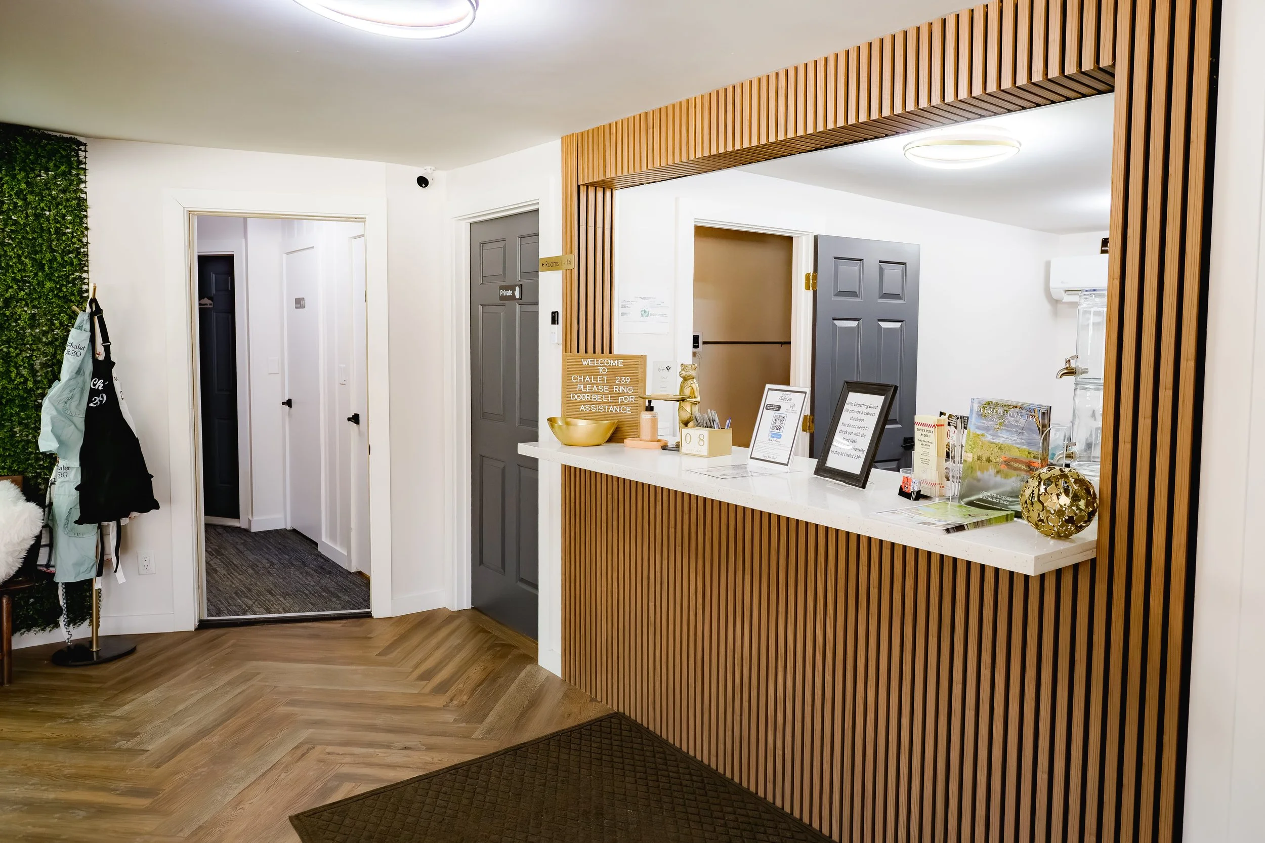 Hotel reception area with a wooden slat partition, a white counter with framed notices, a sign with instructions, a gold bowl, and decorative items. There are hallway doors in the background, a coat rack with bags, and a wall of greenery.