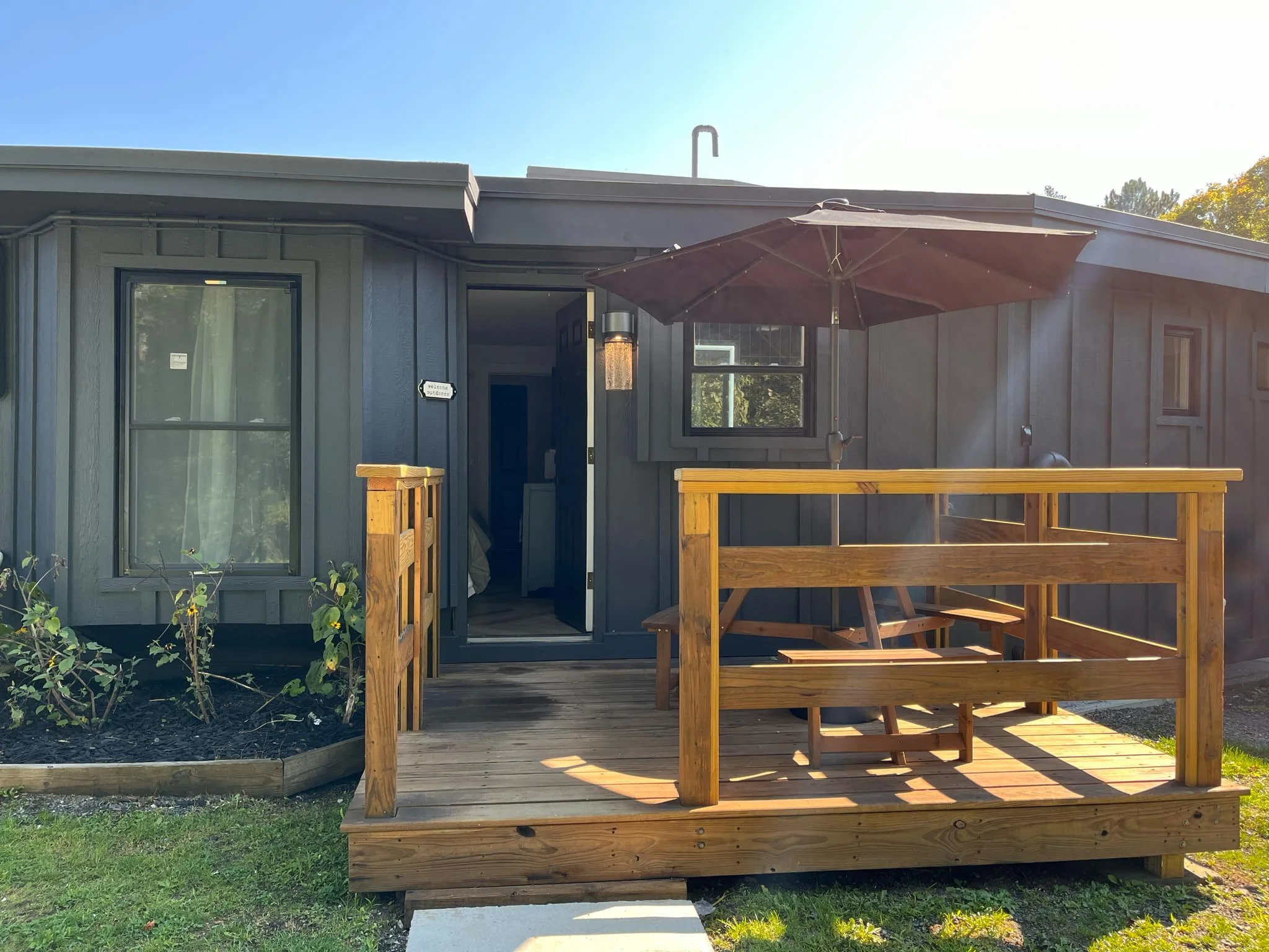 Wooden deck with a picnic table and umbrella outside a dark gray house with two windows and an open door, surrounded by small garden beds and green grass.