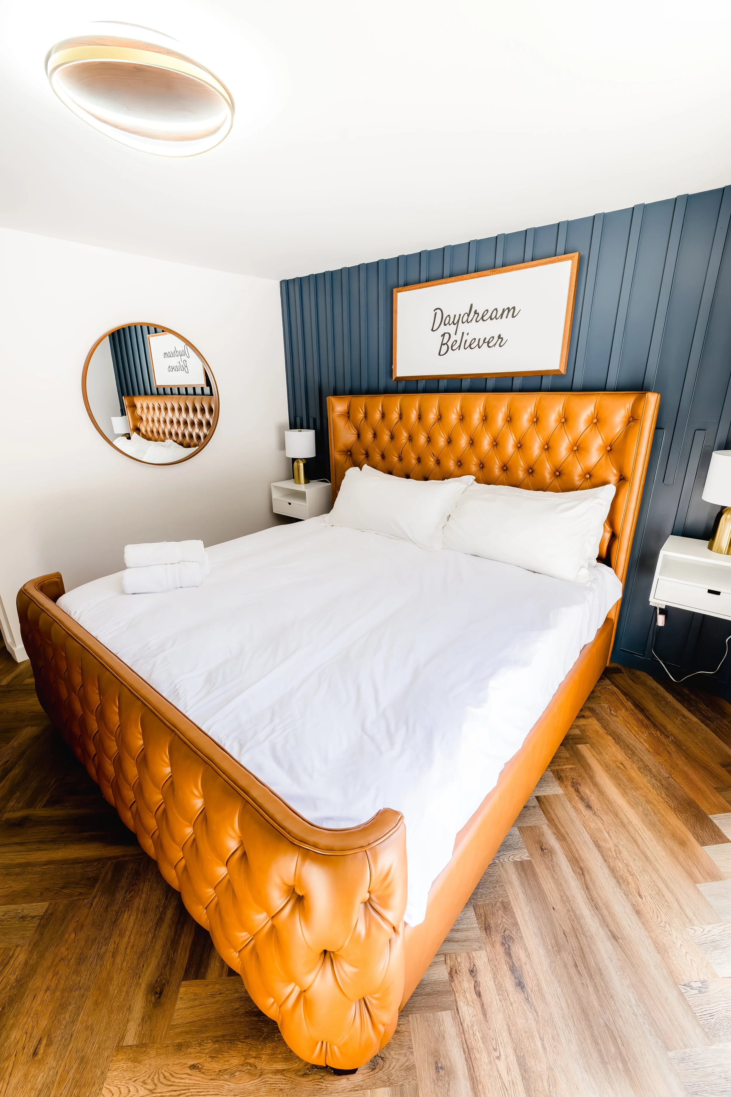 A modern bedroom with a large tufted leather headboard in caramel color, a white bed with pillows and towels, dark wood flooring, two white side tables with lamps, a round mirror on the white wall, and a framed wall art piece that reads 'Daydream Bel