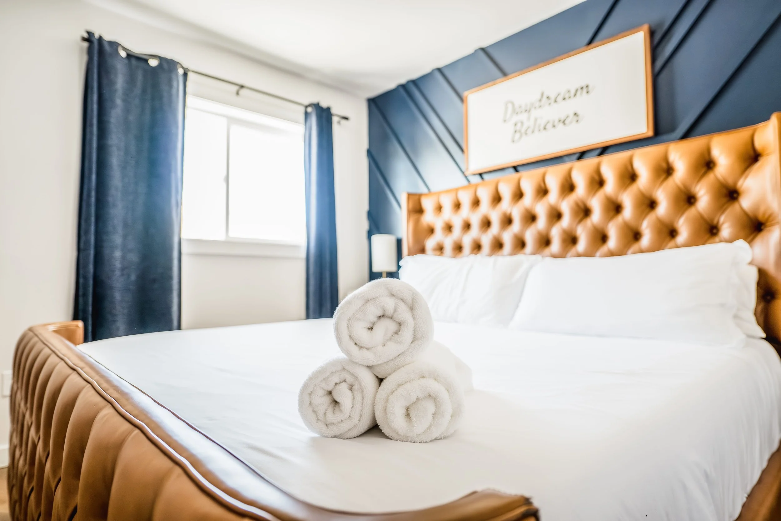 A neatly made bed with a tufted leather headboard, decorated with rolled white towels, in a bedroom with navy blue curtains and a window.