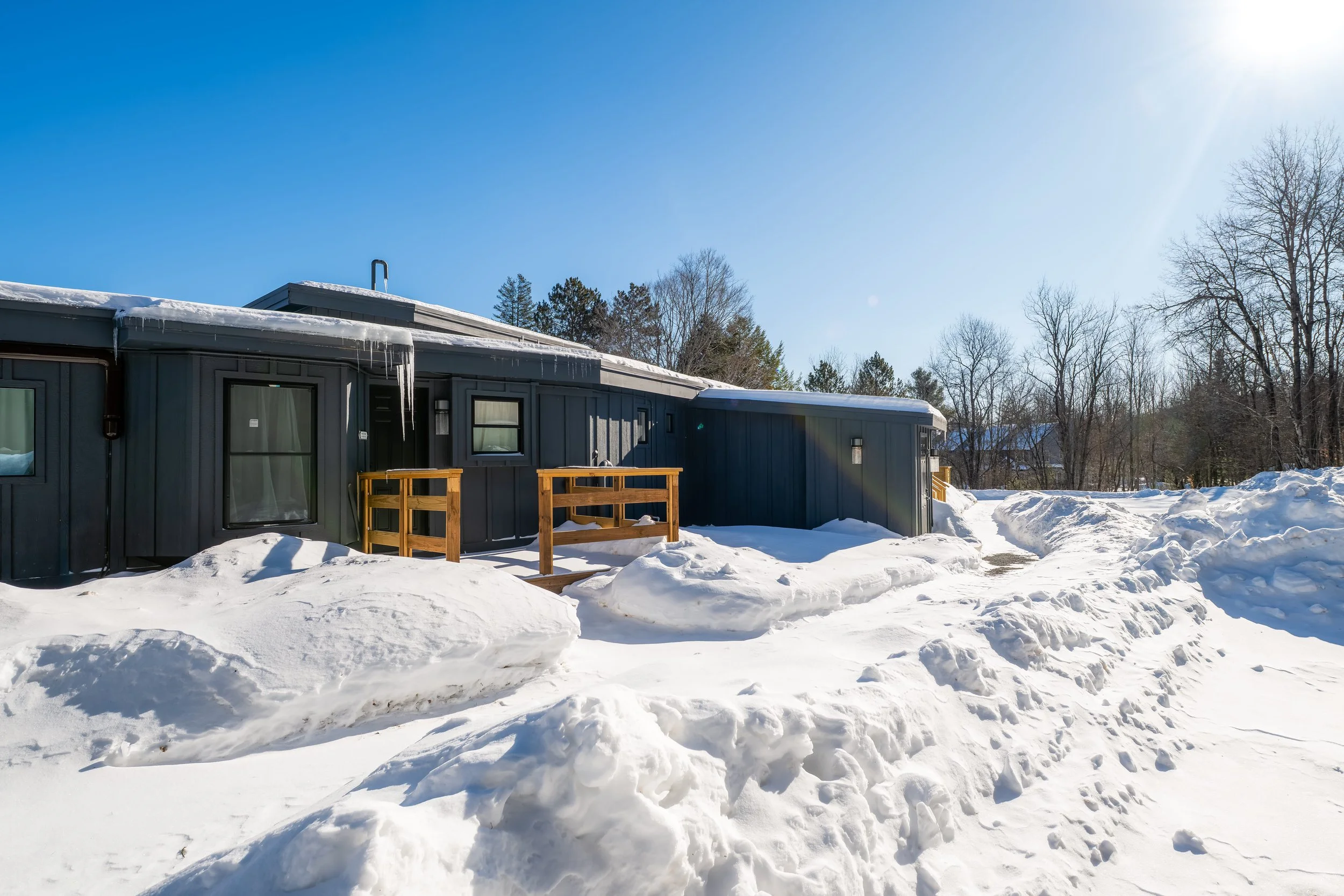 A dark-colored house surrounded by snow with a clear blue sky and leafless trees in the background, bright sunlight, and icicles hanging from the roof.