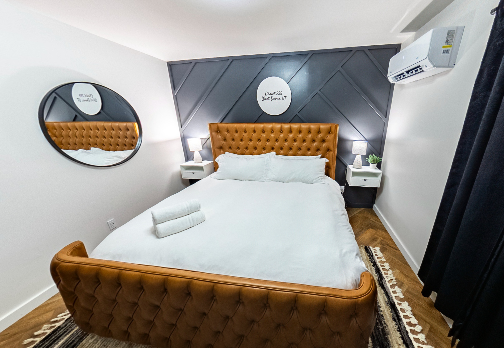A modern hotel bedroom with a tufted leather bed, white bedding, bedside lamps, a mirror, and an air conditioner, decorated with a gray accent wall and a round hotel room sign.
