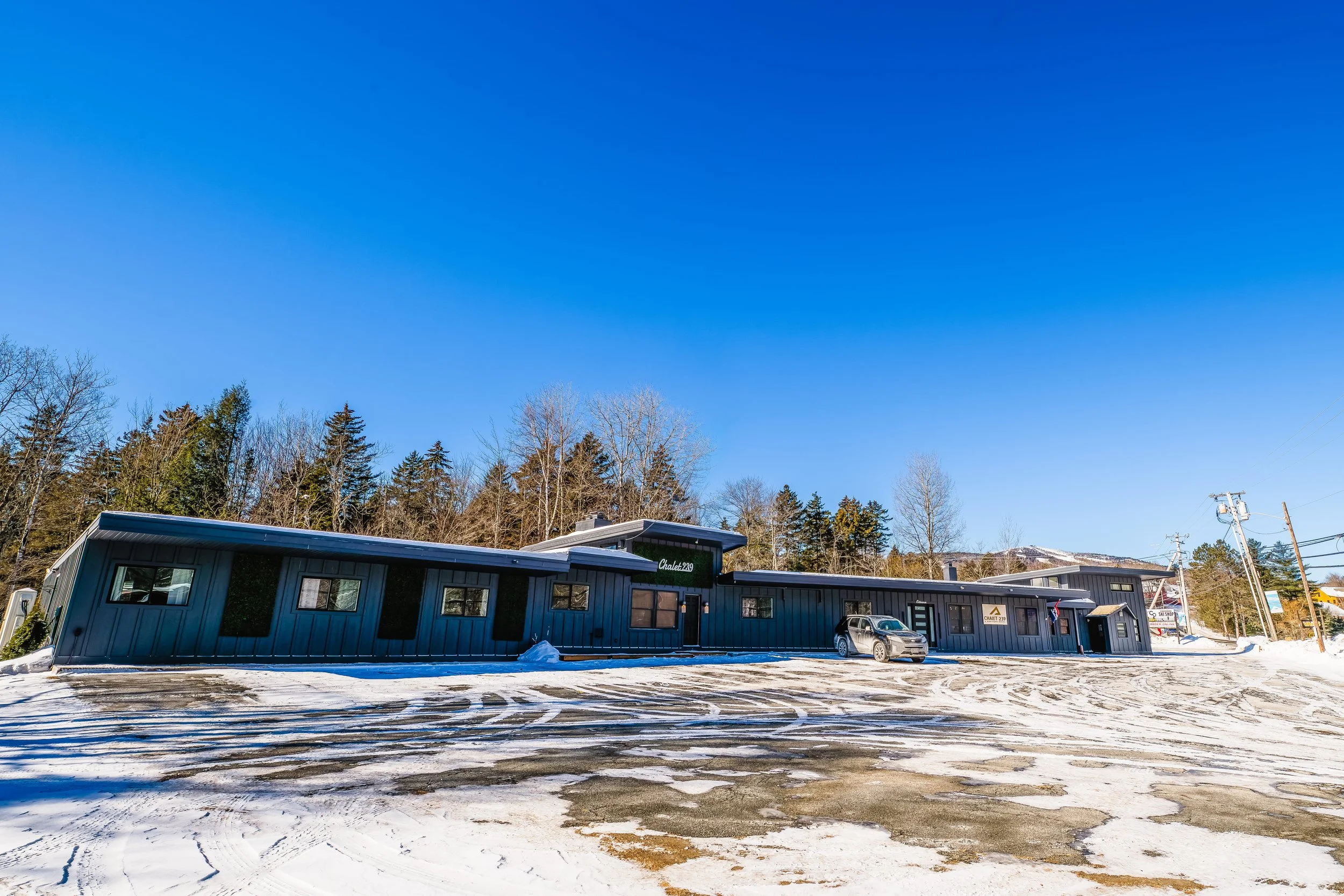 Single-story modern building with large windows, situated on a snowy ground, surrounded by leafless trees and utility poles, under a clear blue sky.