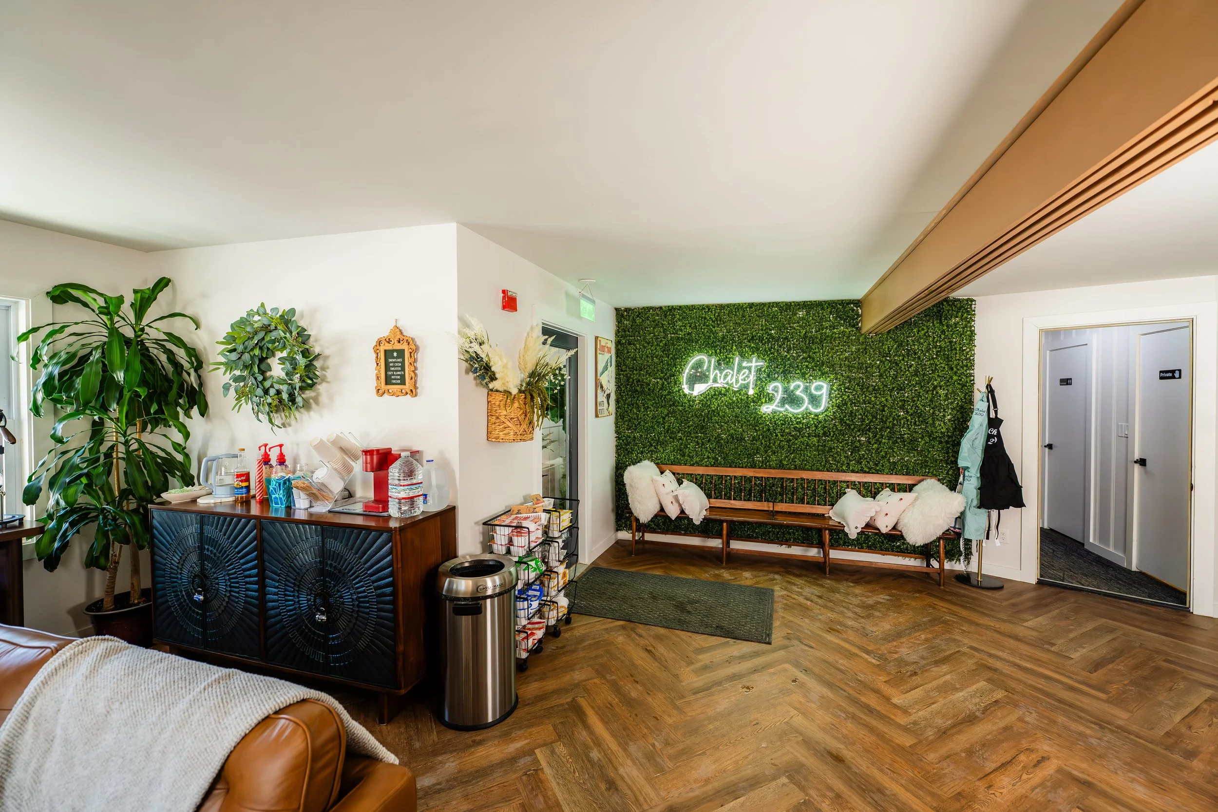 The interior of a cozy waiting area with a green wall featuring a neon sign that says 'Chalet 239'. There are white and pink pillows on a wooden bench, a large potted plant, and a sideboard with water bottles, sanitizer, and other items. The flooring