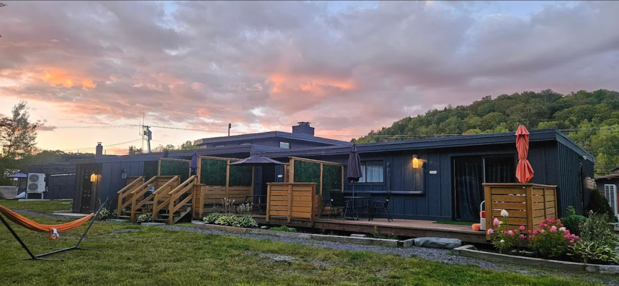 A modern dark blue house with a wooden deck, outdoor furniture, umbrella, and flower beds, set against a sunset with colorful clouds and a green hillside in the background.