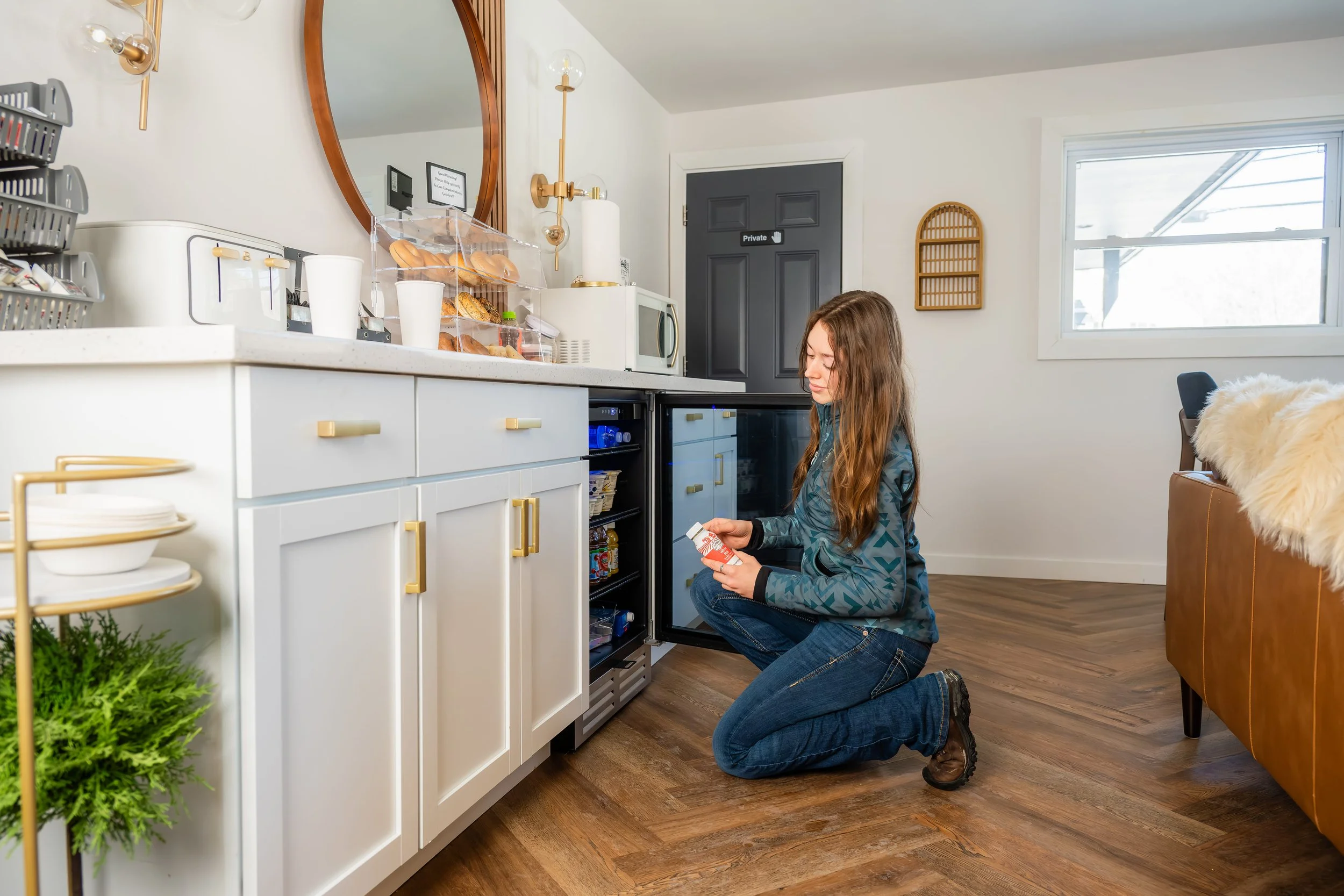 A woman kneeling in front of an open mini refrigerator in a modern kitchen, holding a container of yogurt, with a counter full of breakfast items and a coffee machine nearby.
