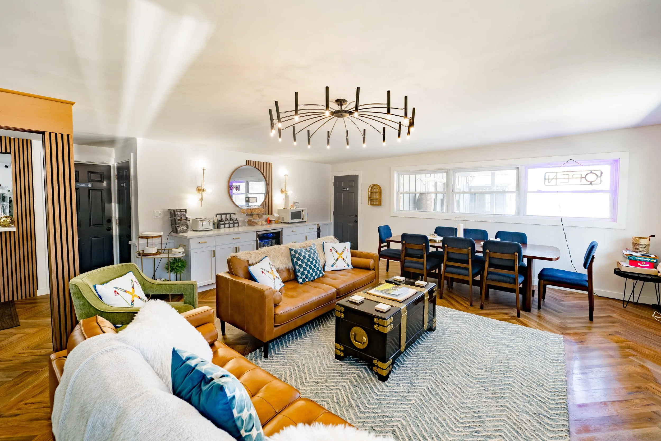Living room with brown and green sofas, a dining table with black chairs, a wooden floor, a modern chandelier, and large windows.