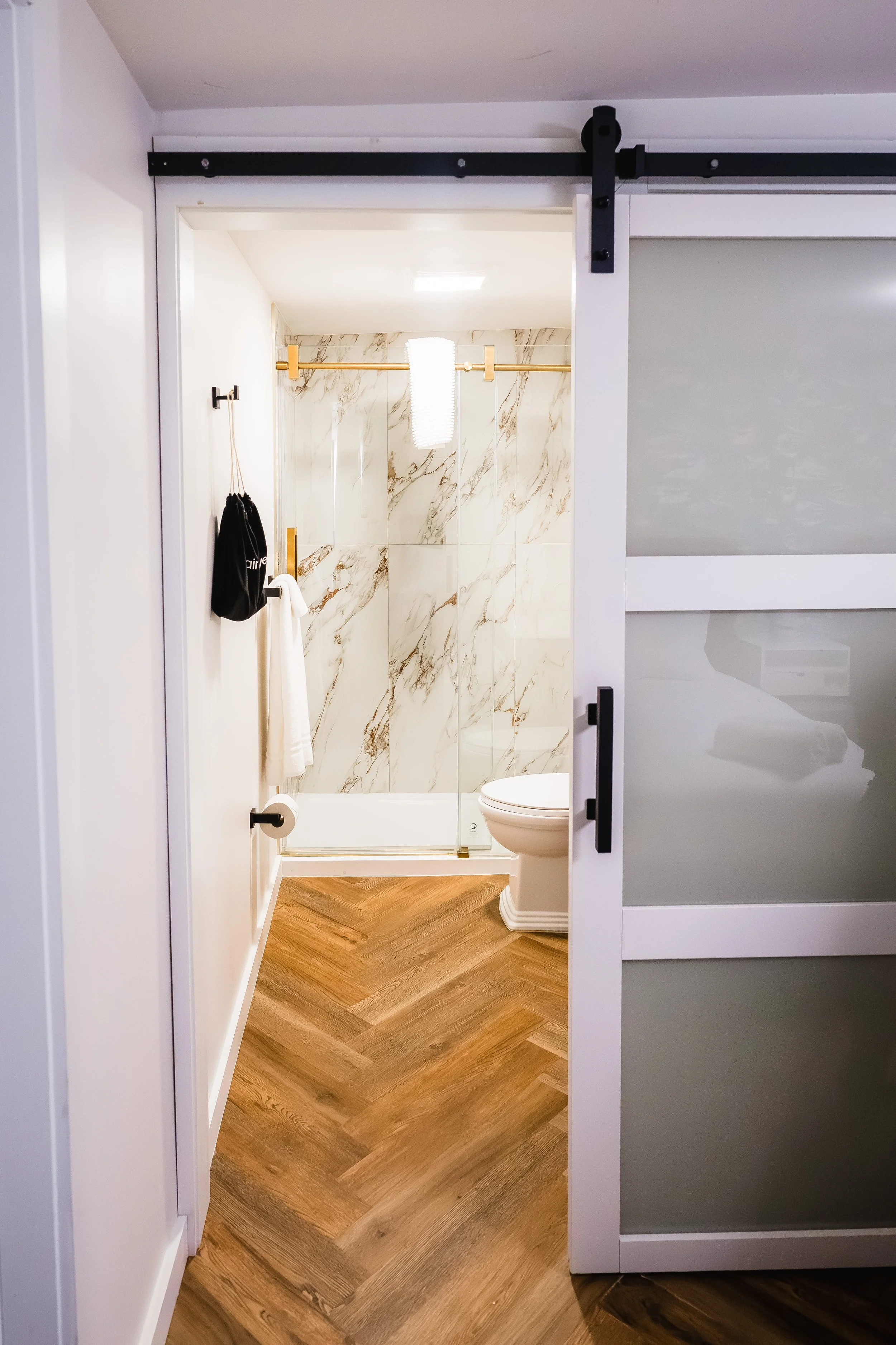 Modern bathroom with a marble shower, toilet, and wood-patterned floor, viewed through a sliding door.