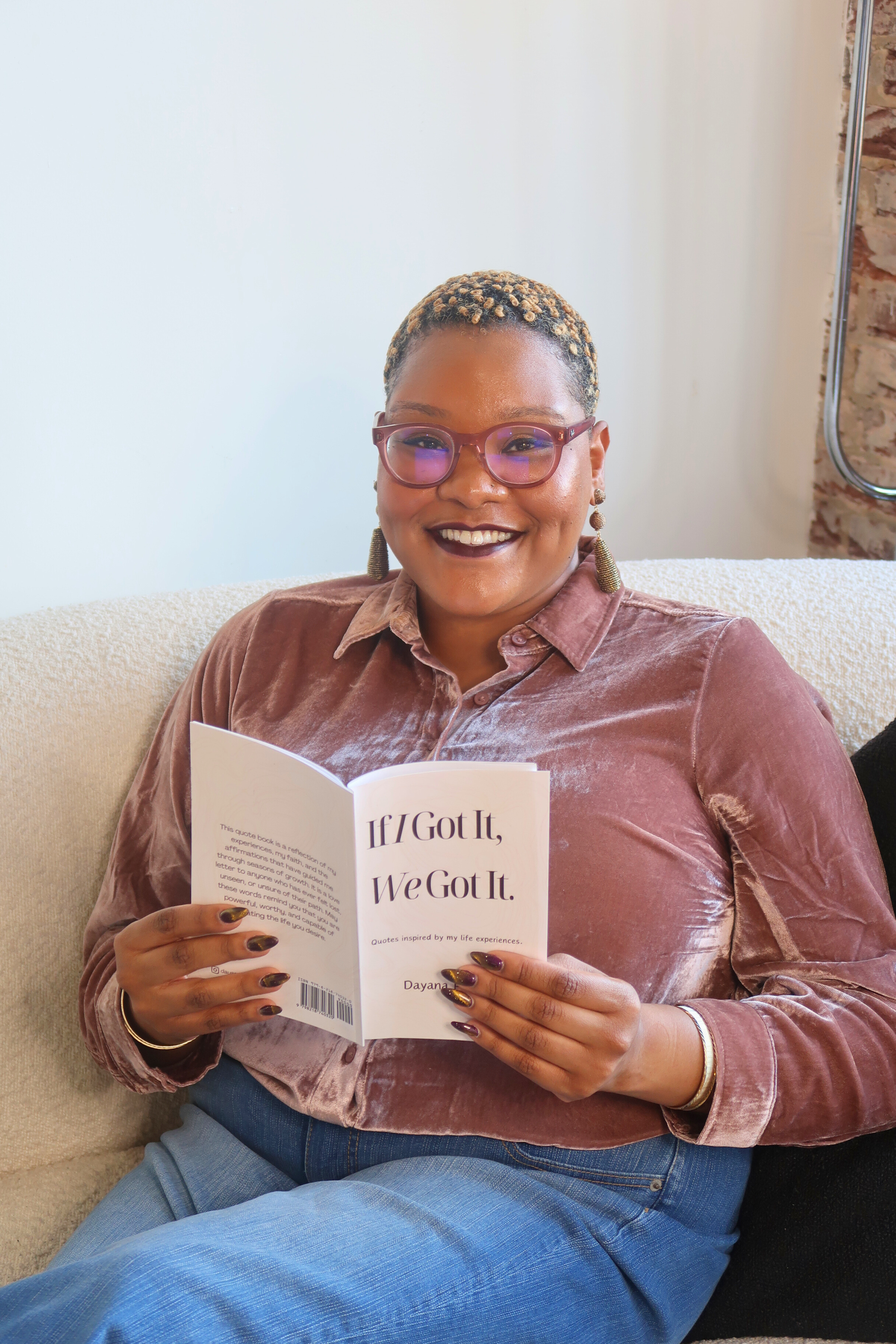 A woman with glasses and earrings sitting on a beige couch, smiling, holding a book open that reads "If I Got It, We Got It."