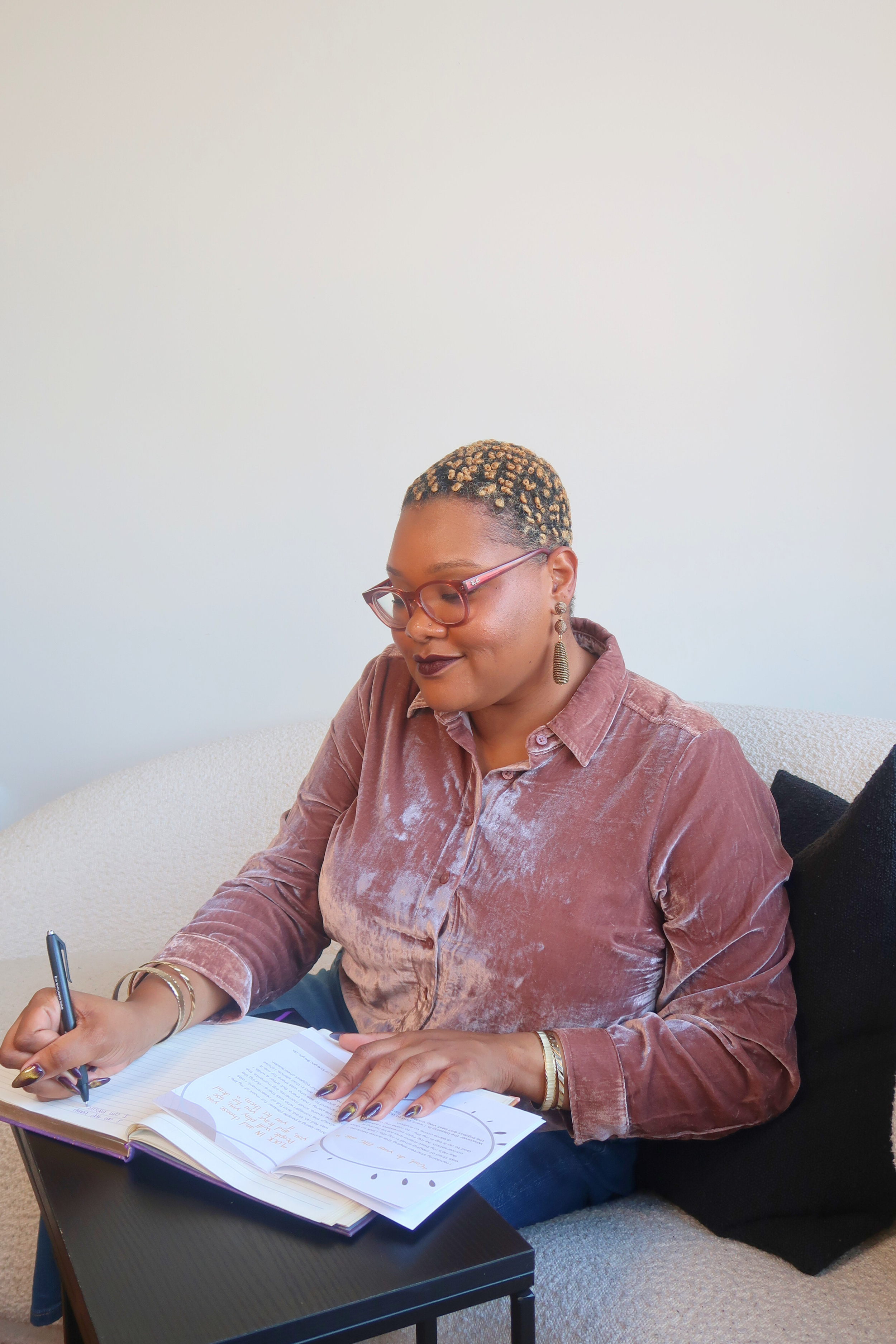 A woman with short blonde curly hair wearing glasses, earrings, and a velvet pink long-sleeve shirt is sitting on a beige couch, writing in a notebook, with a black cushion beside her, in a brightly lit room with a plain white wall.
