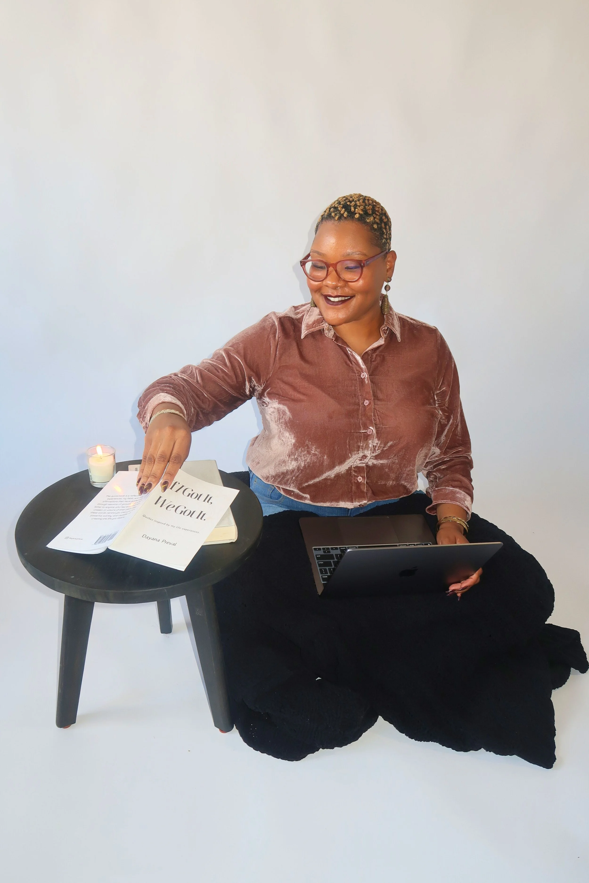 A woman with short, blonde curly hair, glasses, and dark lipstick is sitting on the floor, holding a laptop. She is wearing a velvety brown shirt and black pants. Next to her is a small black table with a candle, a book, and papers, one of which has the title 'If You Got It, We Got It.' She is smiling and appears to be working or studying.