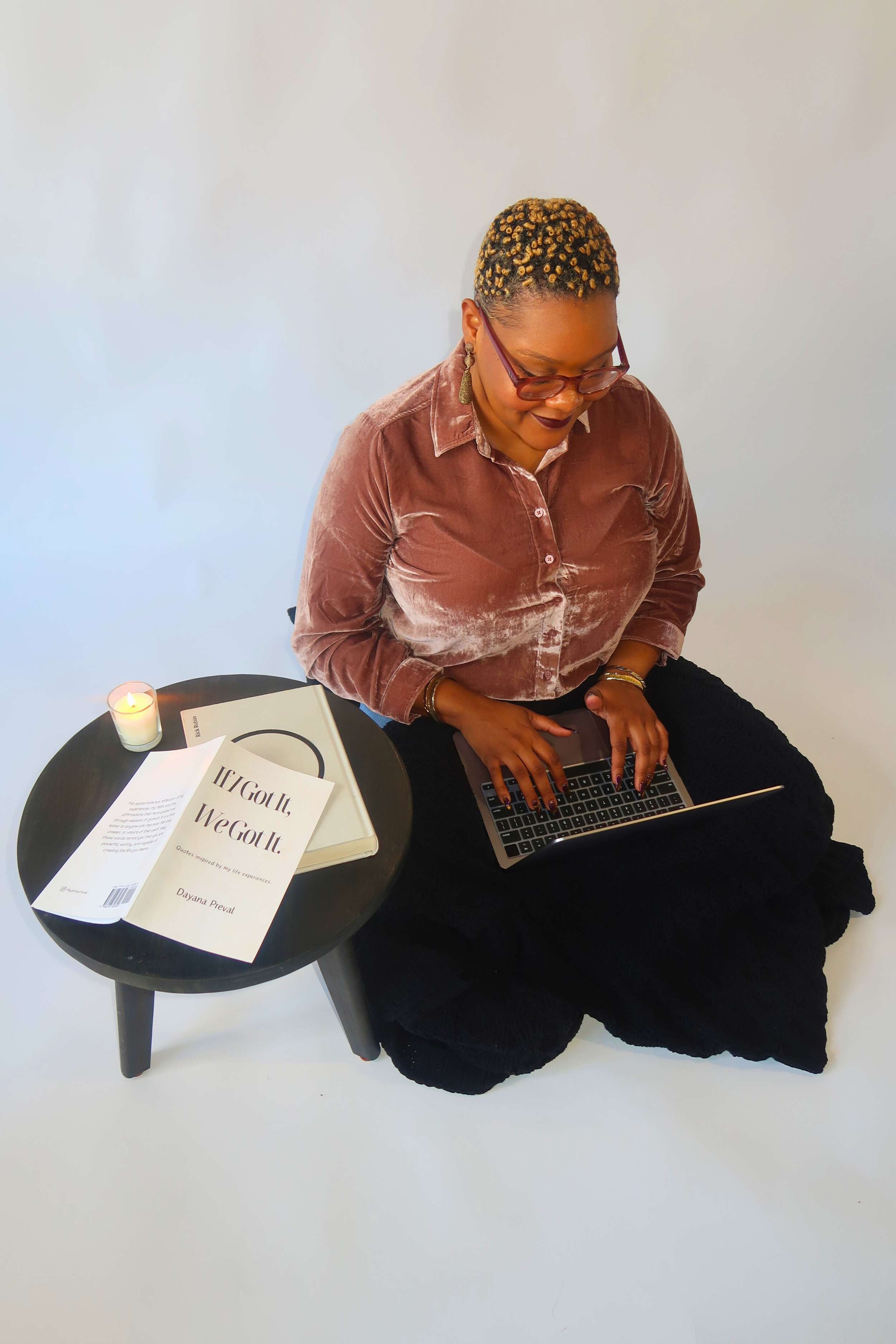 A woman with short, curly hair sitting on a cushion, using a laptop on her lap. Beside her is a small black table with a lit candle, a book, and a printed quote saying 'If I Got It, We Got It.'