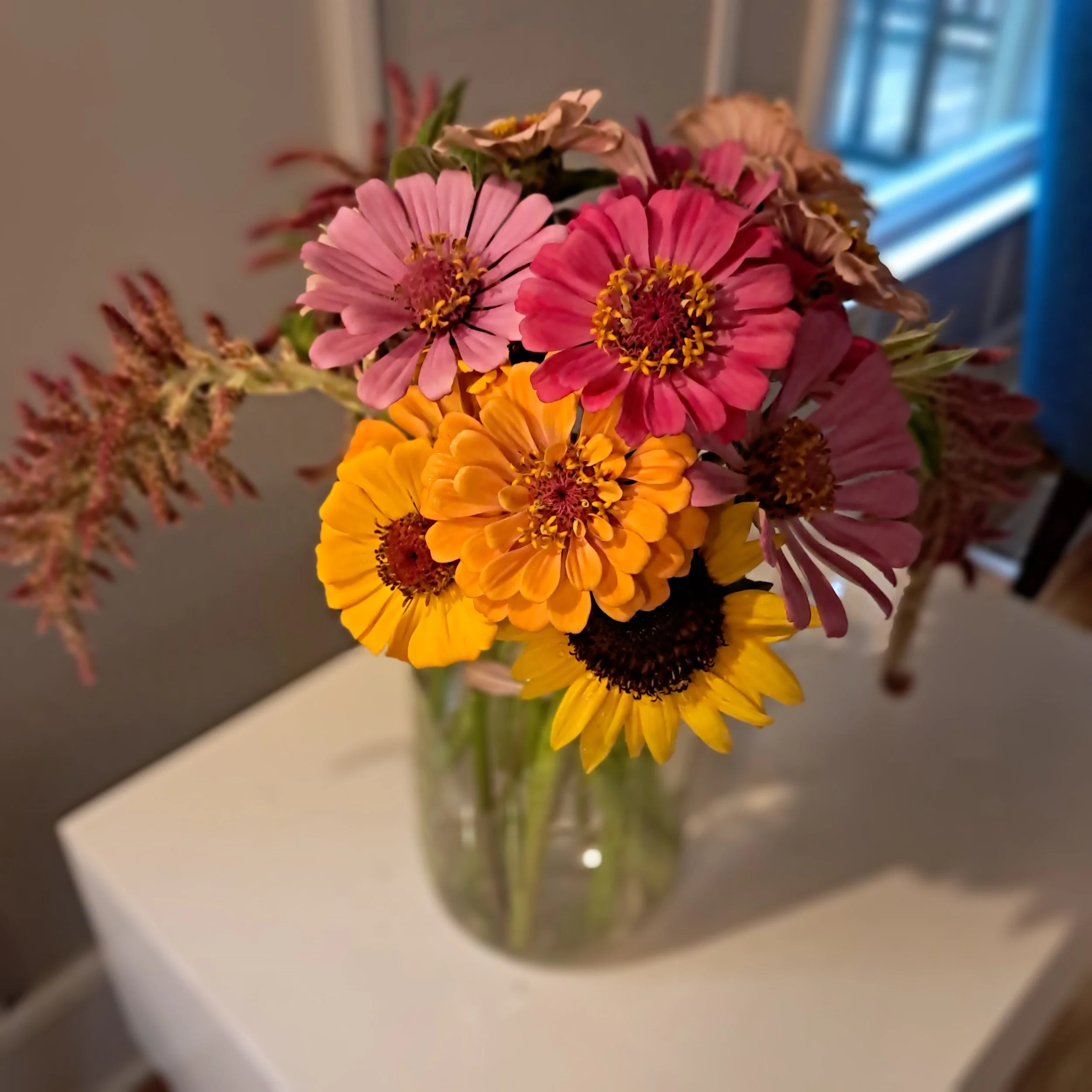 A bouquet of colorful flowers including pink, red, yellow, and orange blossoms in a clear glass vase on a white table, with a blurred background of a room with a window.