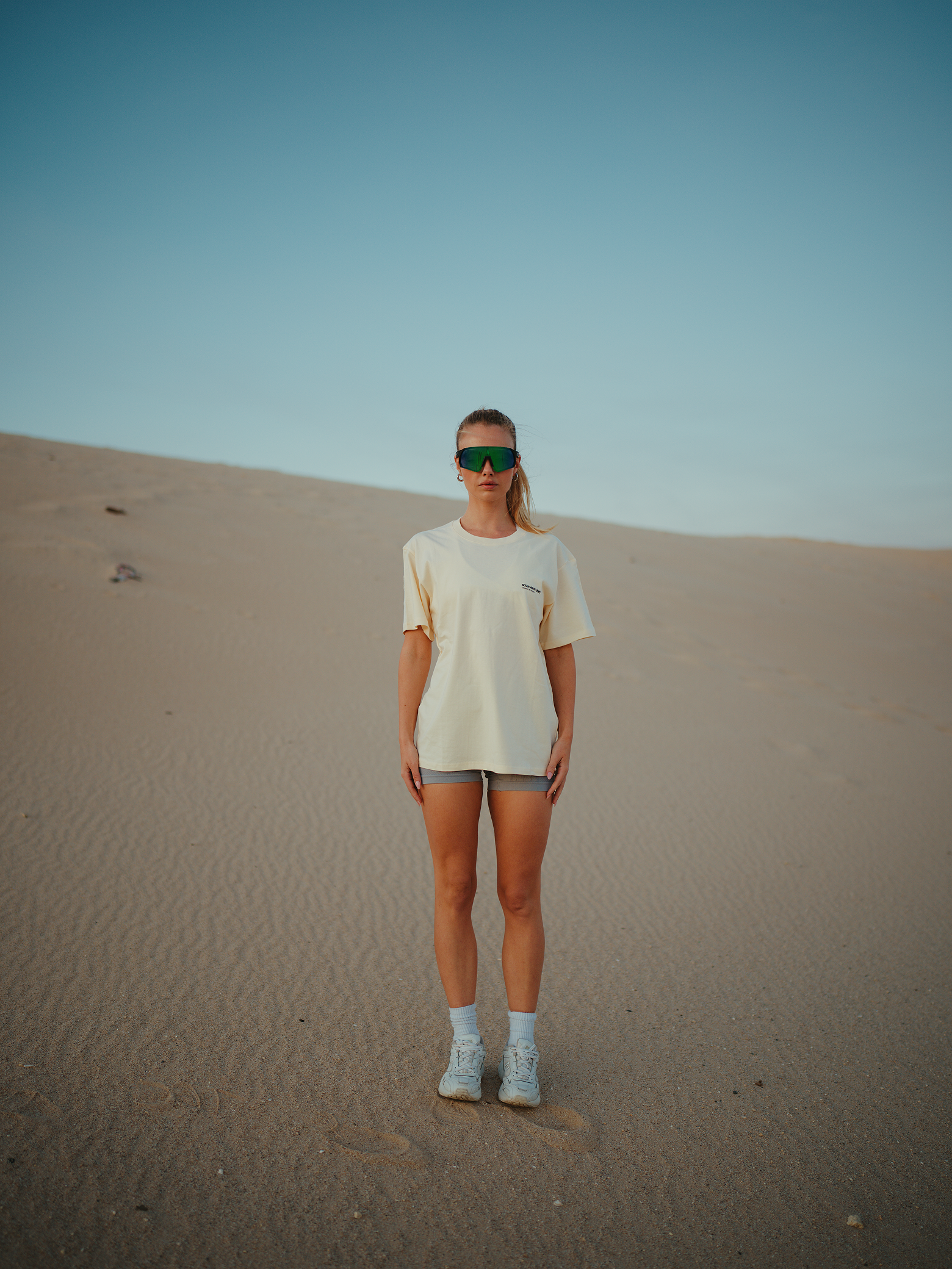 A woman standing in a sandy desert landscape wearing sunglasses, a cream-colored oversized t-shirt, gray shorts, white sneakers, and white socks, looking directly at the camera.