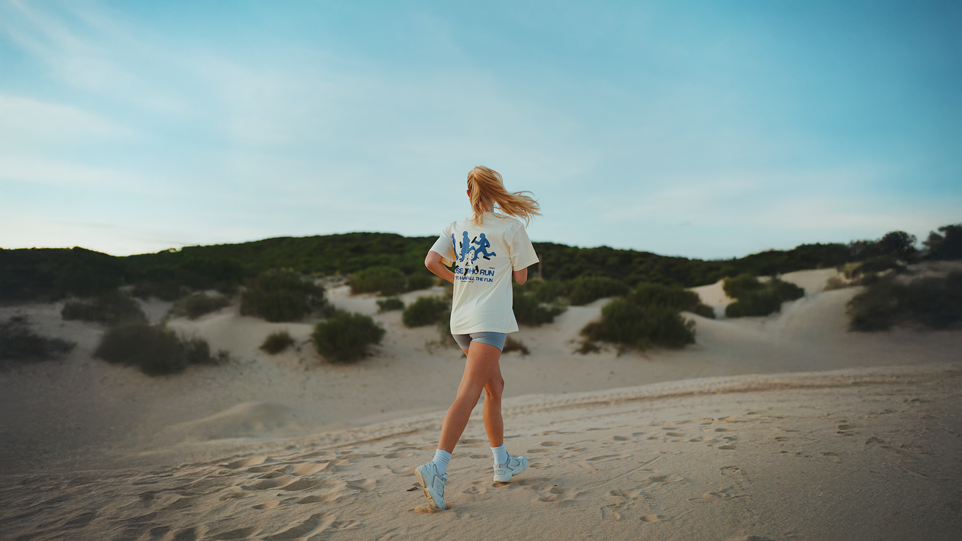 A woman jogging on a sandy beach with dunes and green bushes under a clear blue sky.