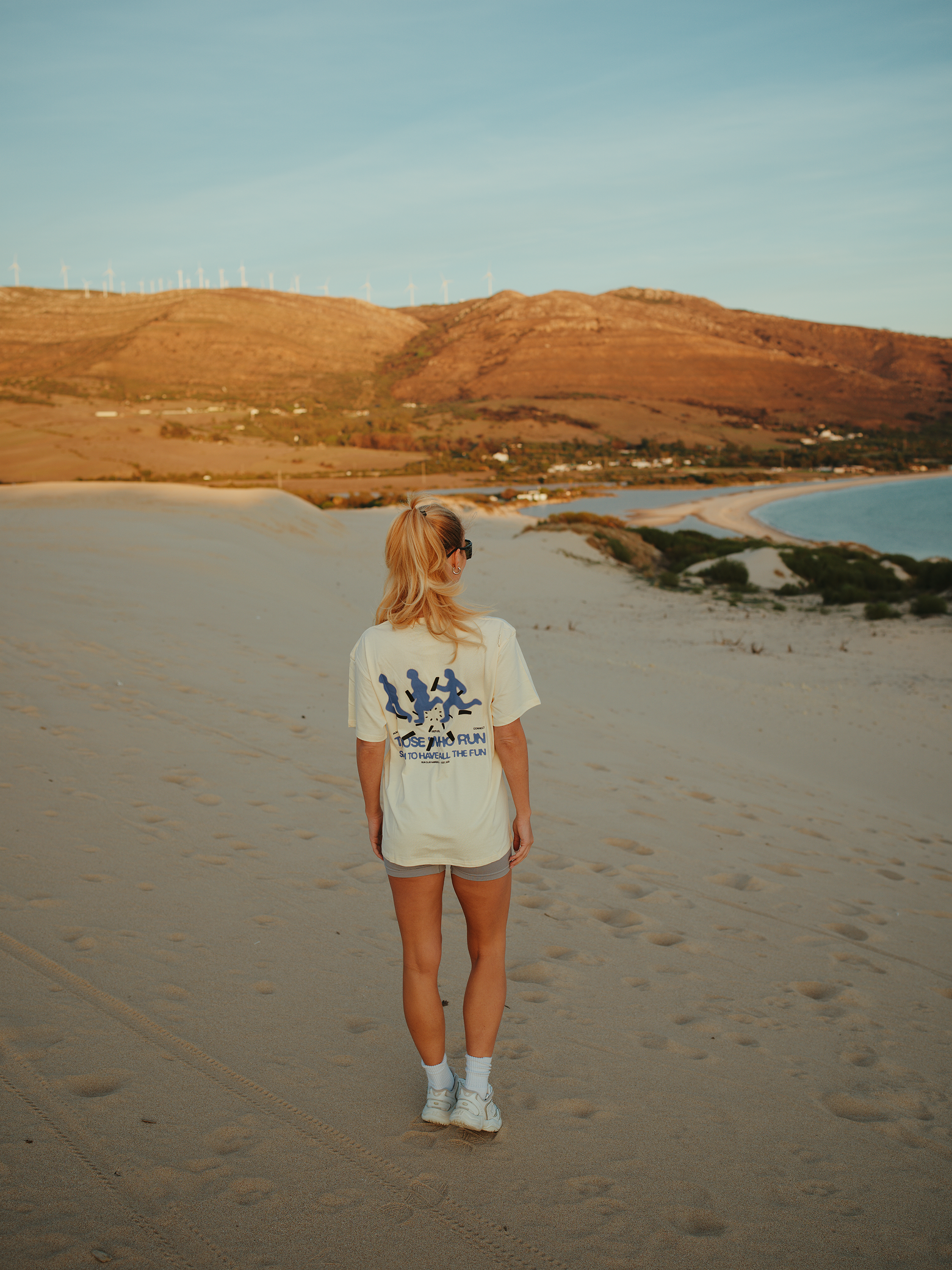 A woman with blonde hair in a ponytail wearing sunglasses, a white T-shirt with blue graphics, shorts, white socks, and sneakers walking on a sandy beach with hills, wind turbines, and the ocean in the background during sunset.